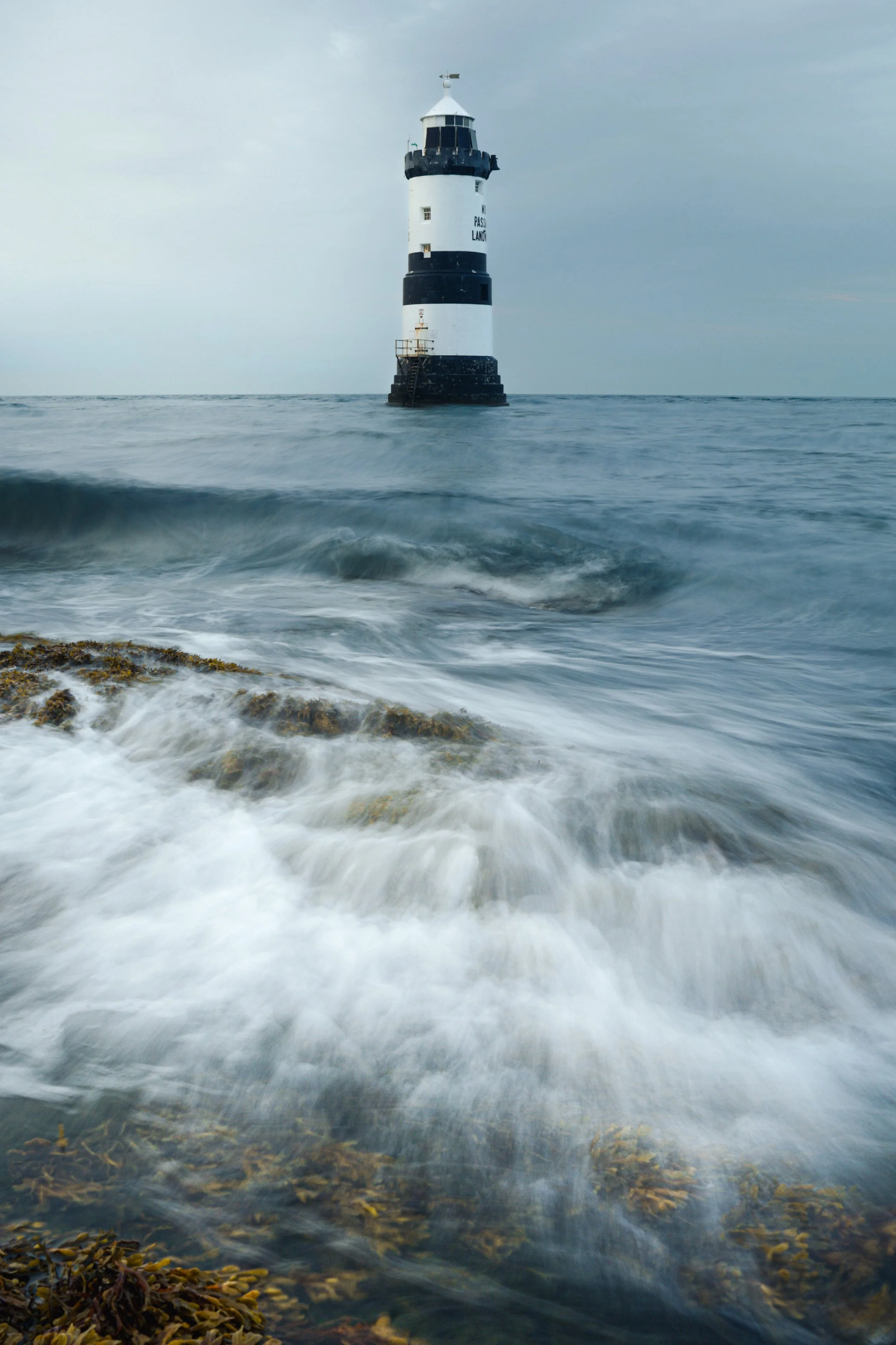 A lighthouse standing amid ocean waves with seaweed in the foreground and cloudy sky in the background.