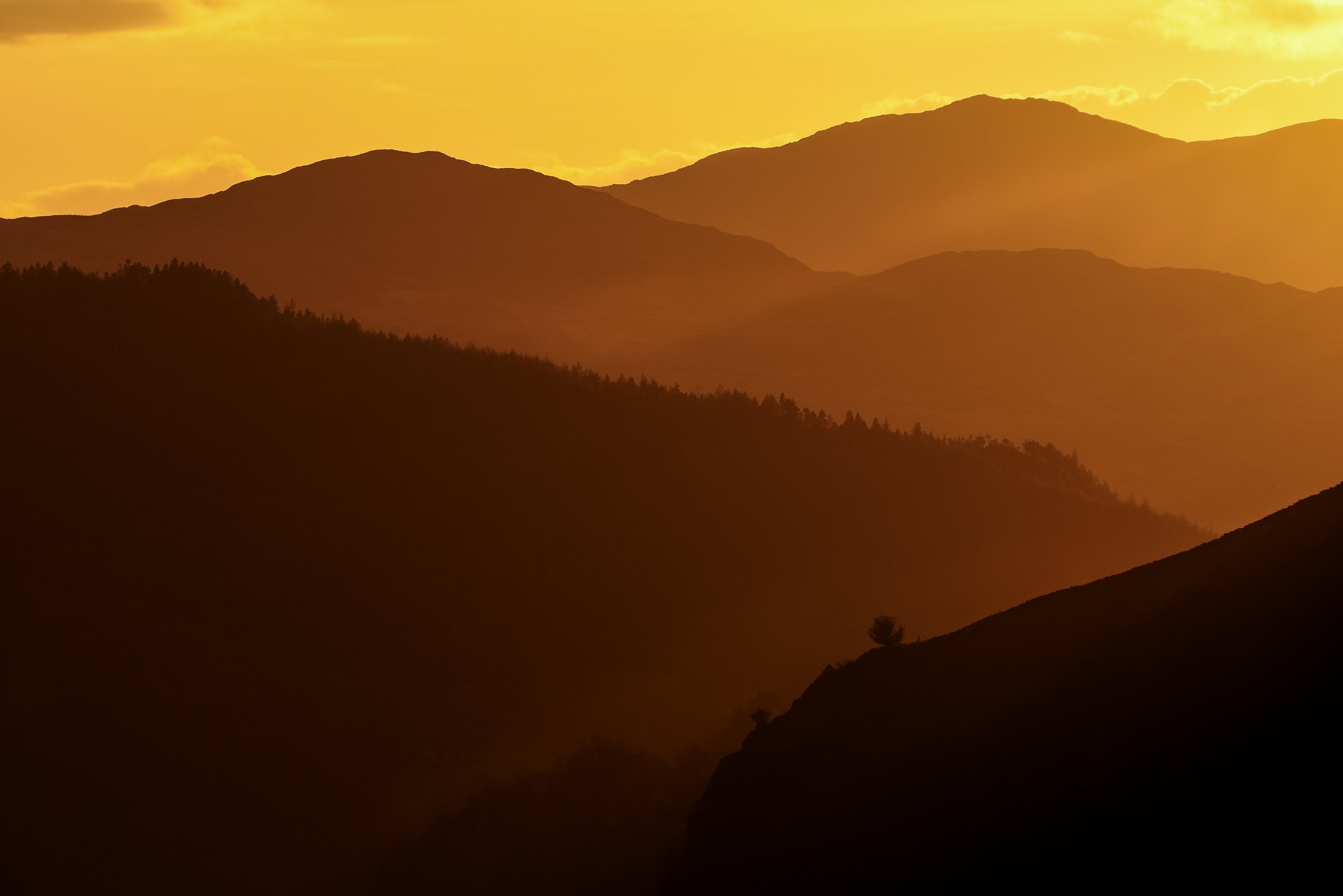 Silhouetted mountain range at sunset with layers of hills fading into the distance and a warm golden-orange sky.