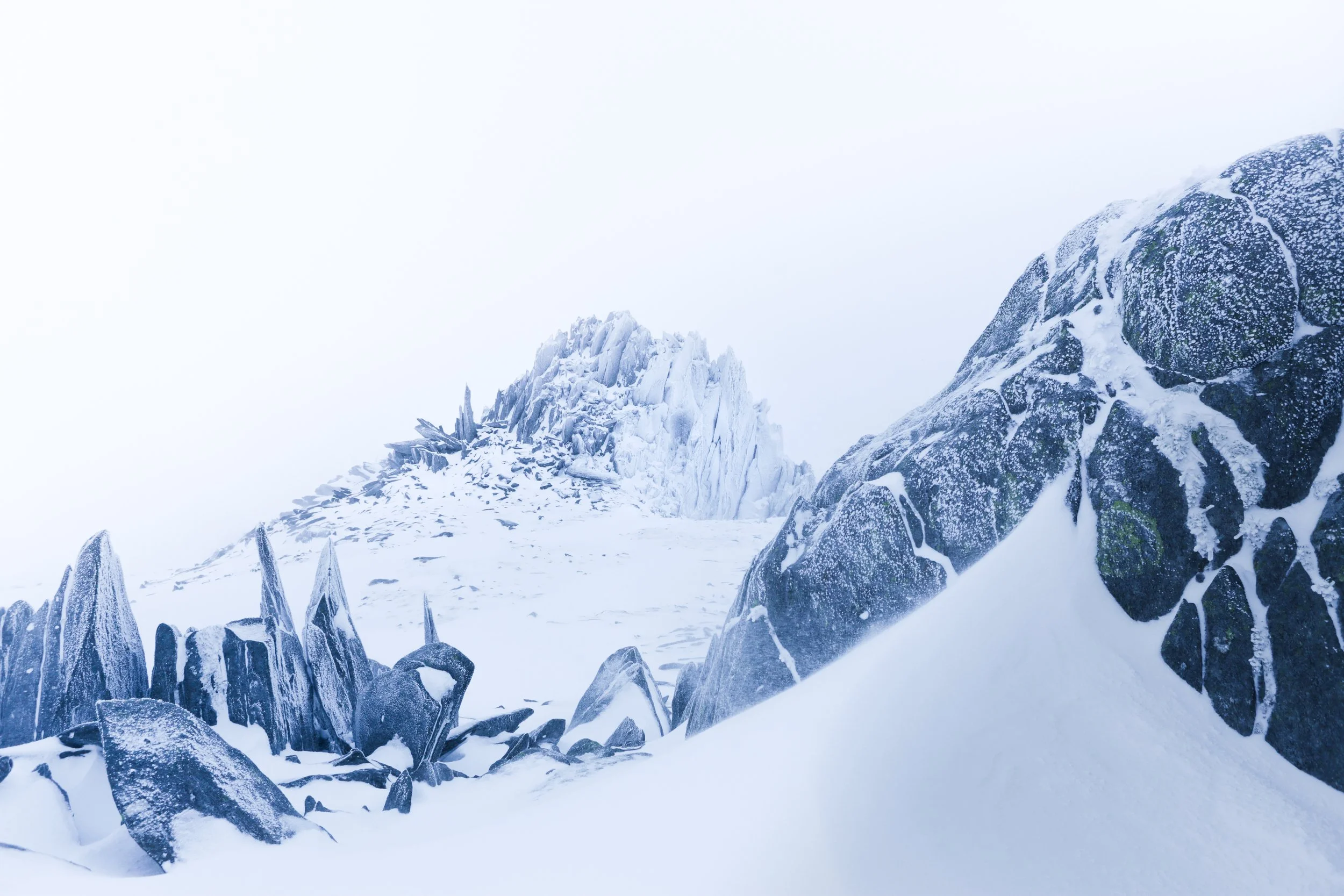 Snow-covered rocky mountain landscape with large boulders and a jagged icy peak in the background, foggy sky.