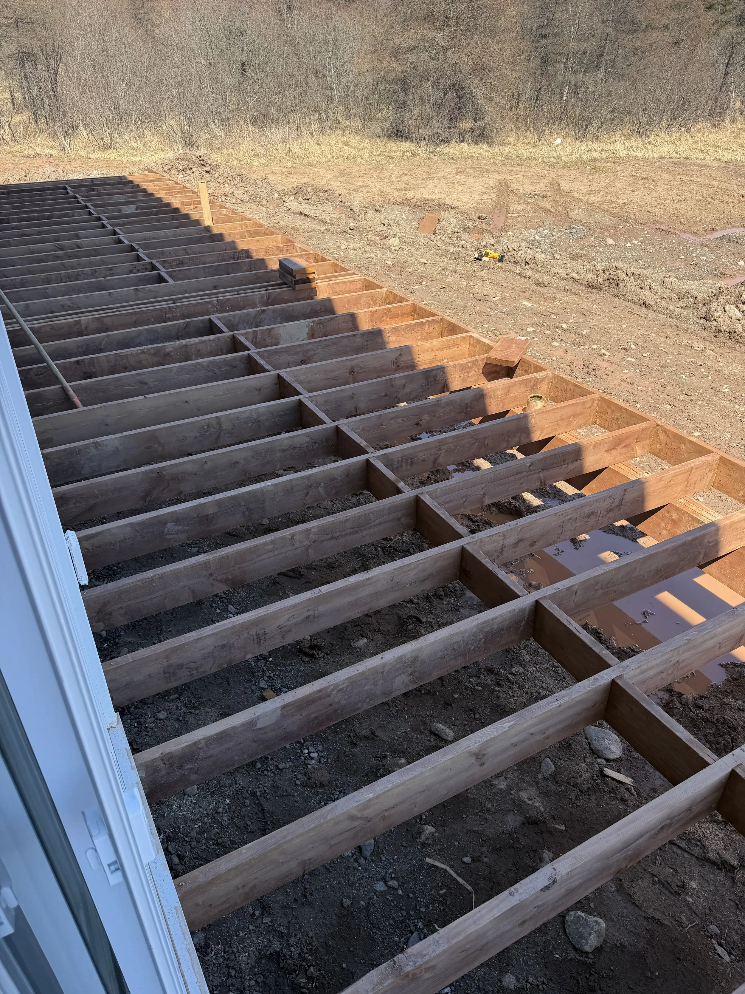 Unfinished wooden deck framing on a construction site with dirt ground and leafless trees in the background.