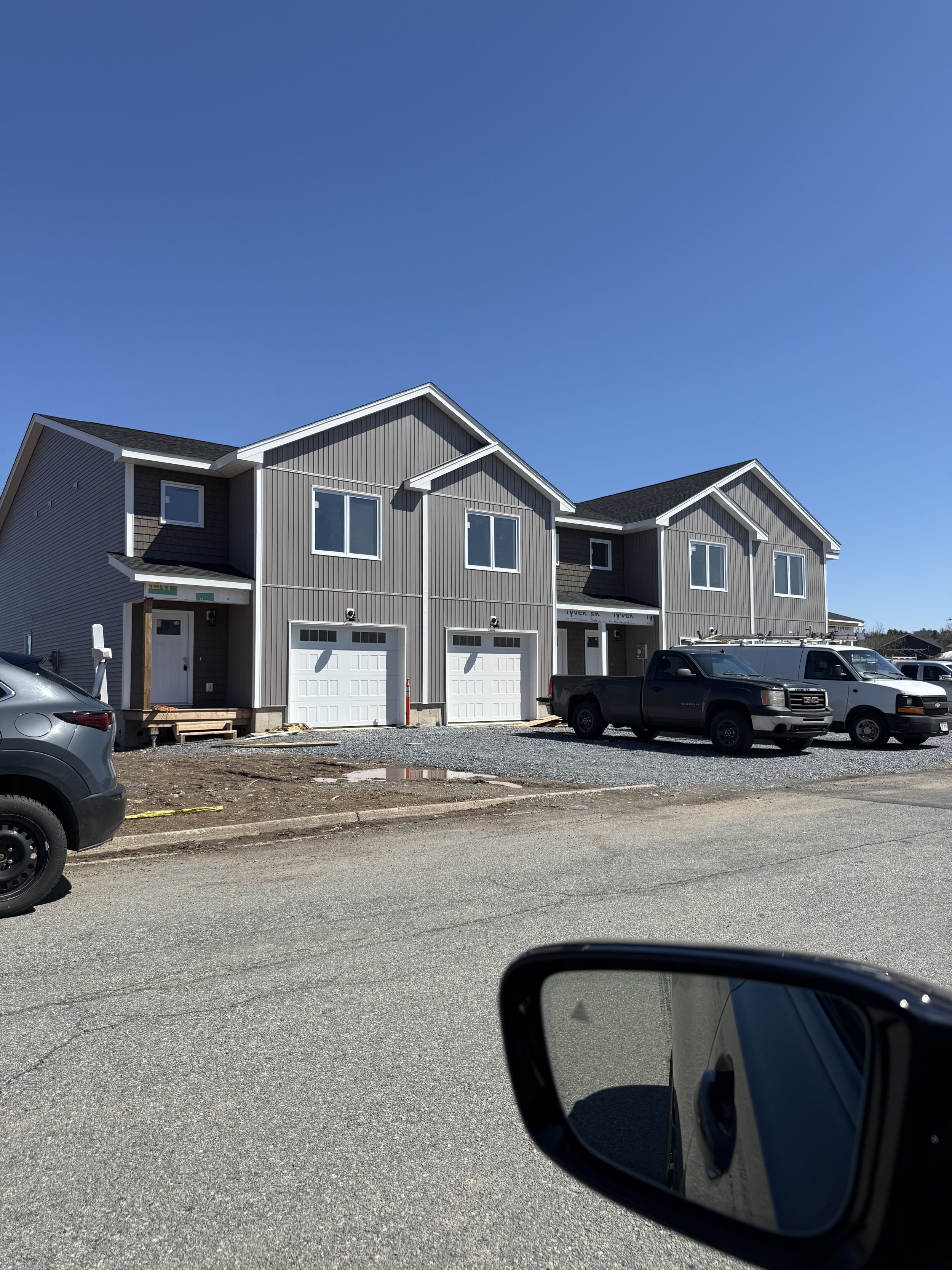 Photo of a row of new townhouses with gray siding and white garage doors under a clear blue sky. There are vehicles parked in front, and some construction materials are visible near the houses.