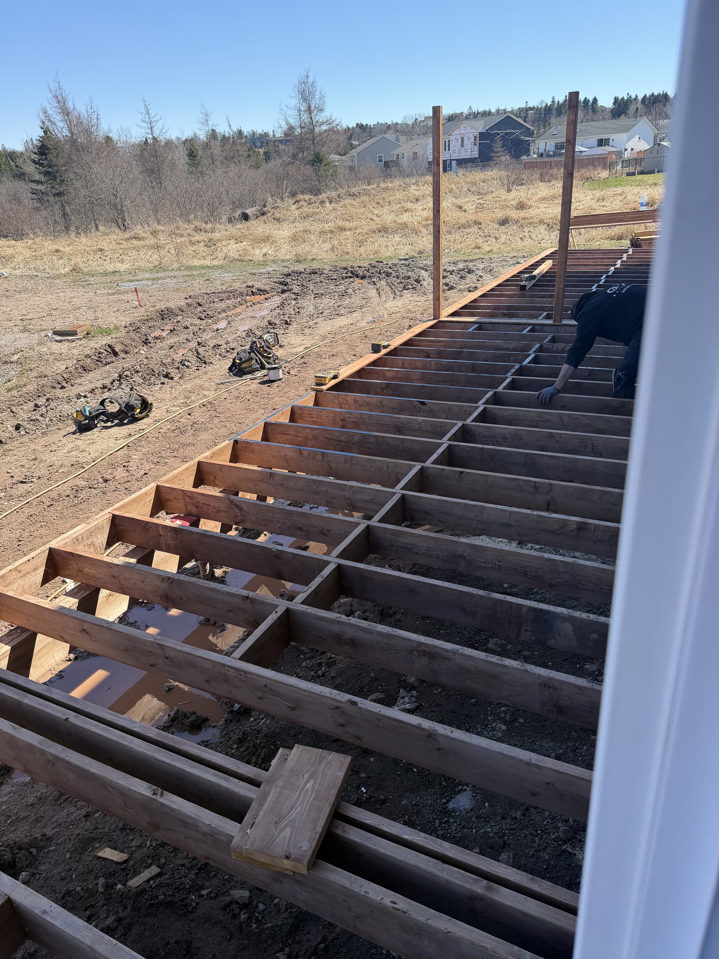 Construction site showing wooden framework for a deck or porch, with tools and construction materials scattered around, and a worker kneeling and working on the structure.