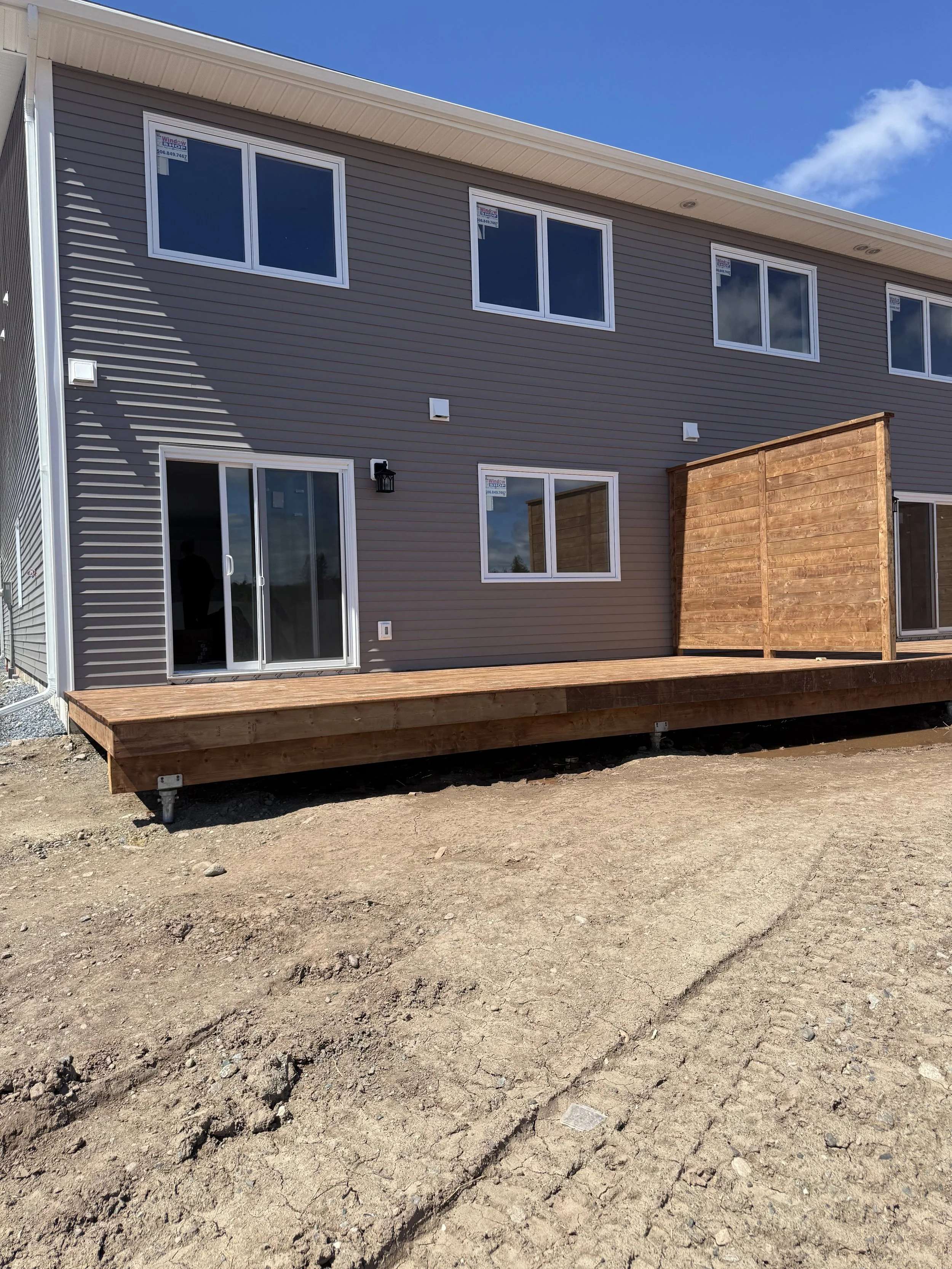 Newly constructed house in Saint John, NB with large windows, a sliding glass door, a wooden deck, and a wooden privacy fence, under a clear blue sky.