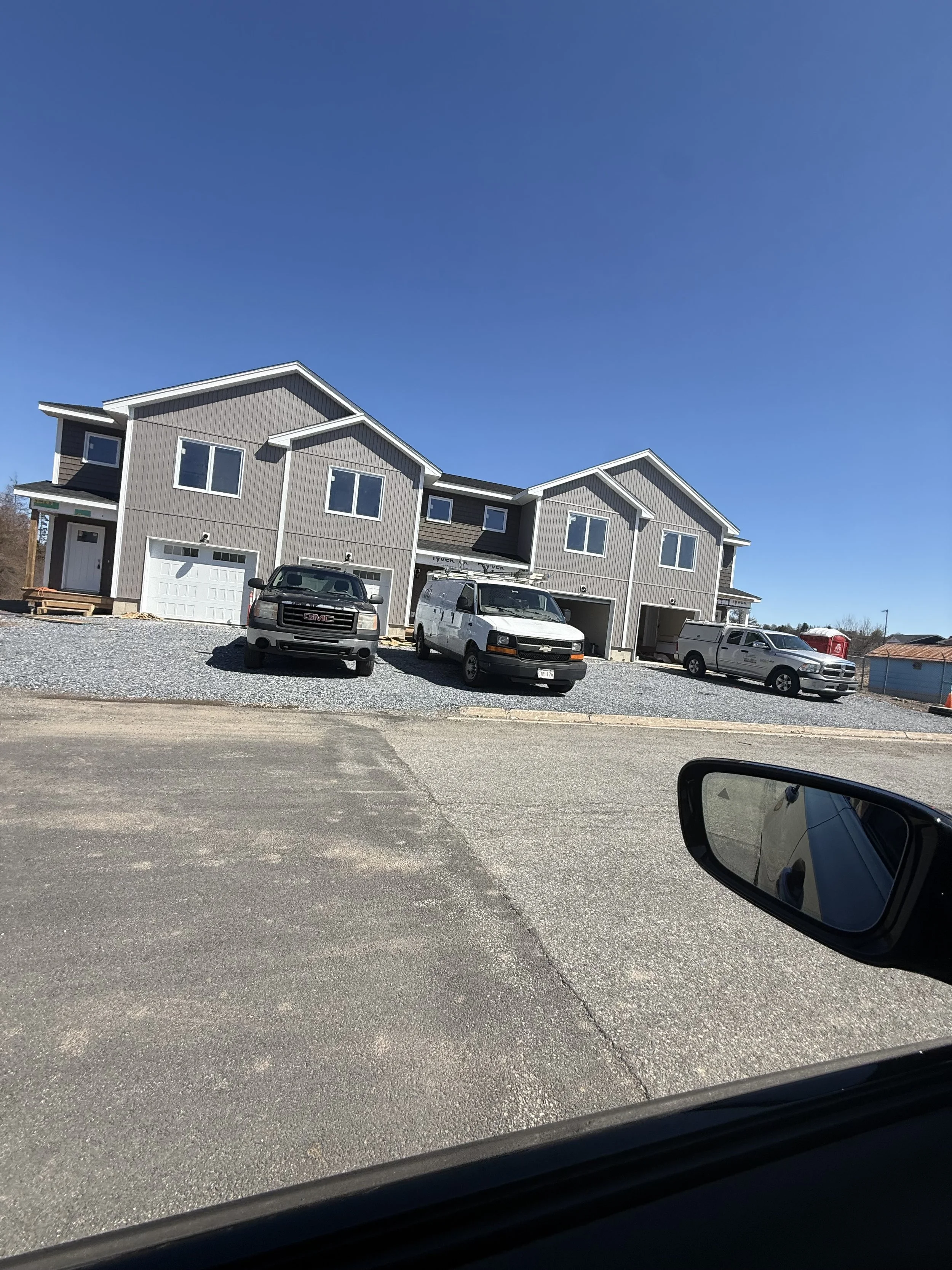 View of newly constructed townhouses with gray siding, white trim, and attached garages; there are multiple vehicles parked in front, including a GMC truck and a white van, under a clear blue sky.