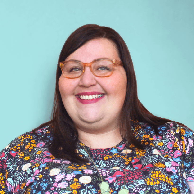 Headshot of a fat white woman smiling. She has peach glasses, long dark hair, and is wearing a flowered shirt.