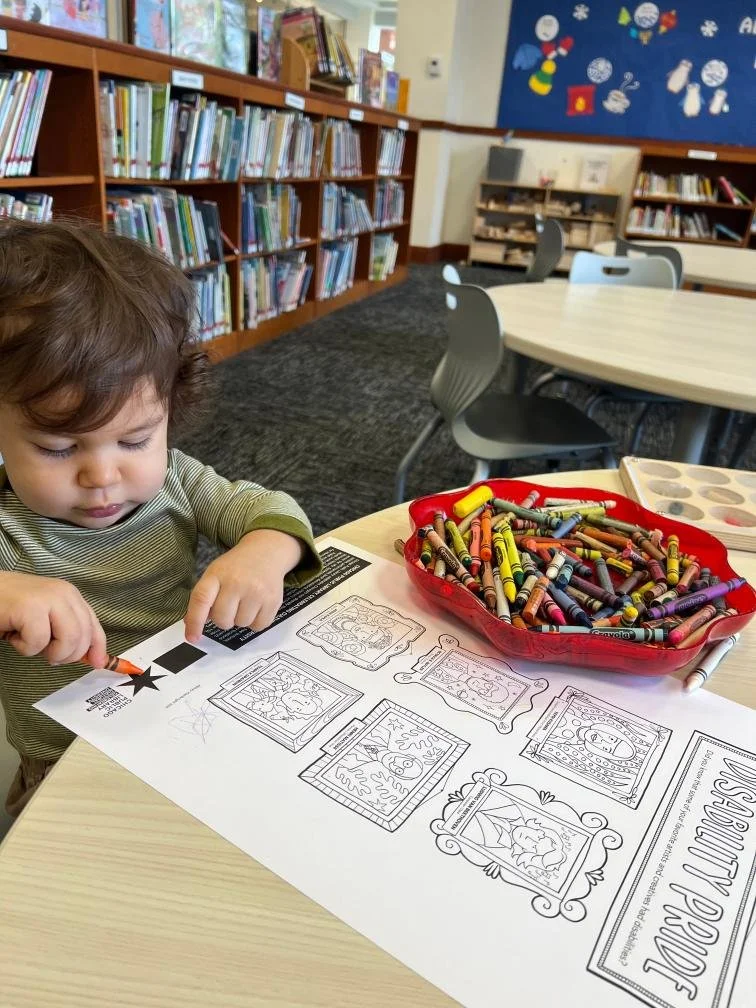A toddler draws with a crayon on the Disability Pride coloring sheet at a table at a library