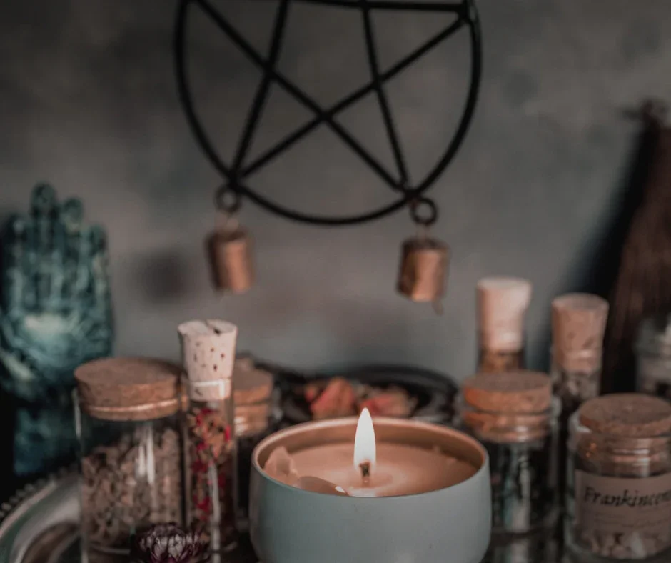 A lit candle in front of a collection of jars and containers on a surface, with a dark wall and a pentagram decoration hanging above.