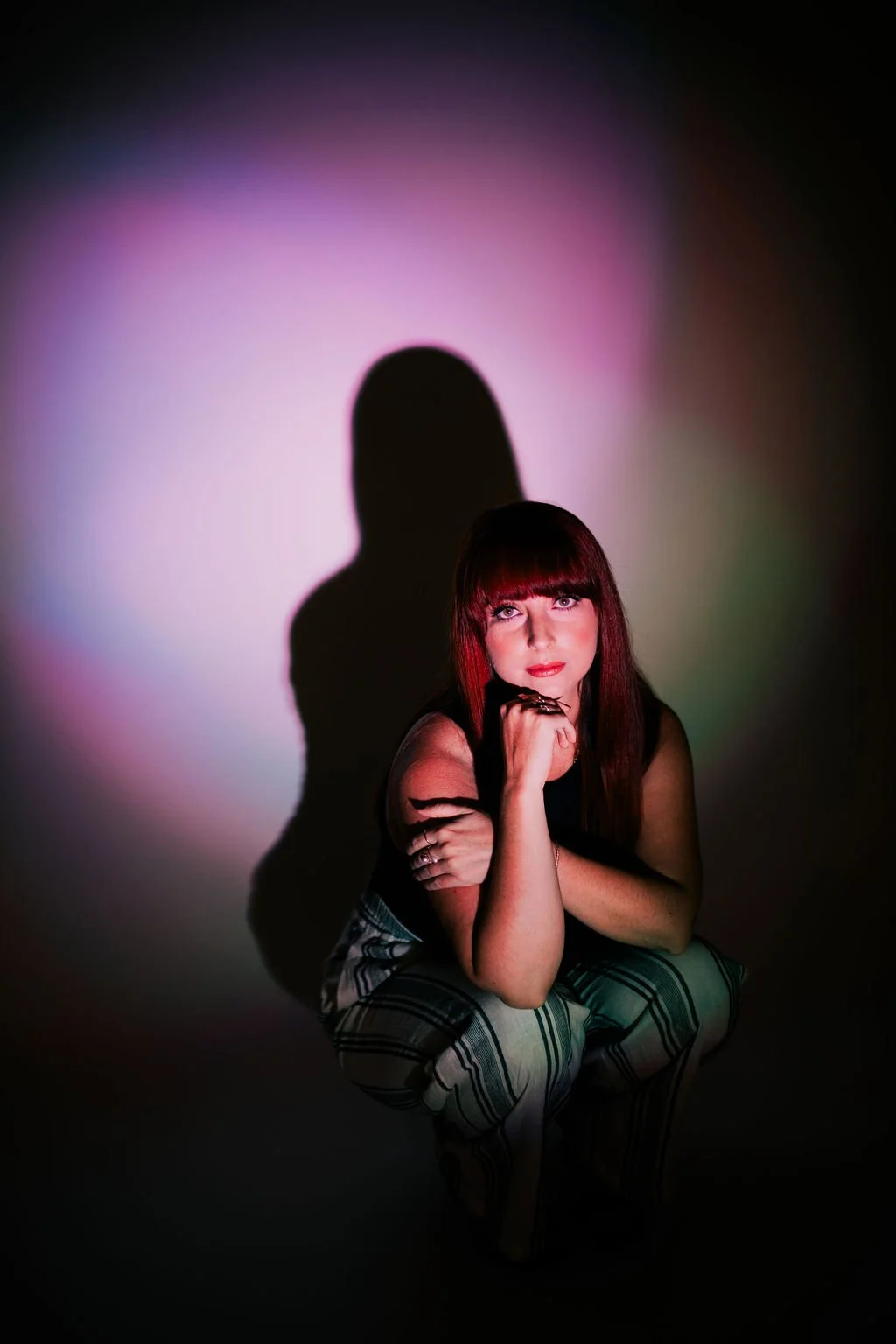 A woman with long red hair and bangs crouches against a wall illuminated by multicolored lights. Her eyes gaze softly at the camera, and she rests her chin on her right hand while her left arms are crossed over her knees. The colorful pastel lighting creates a shadow on the wall behind her.