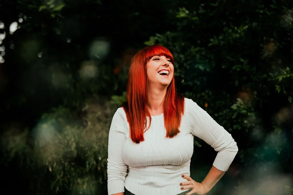 A woman with bright red hair and a white long-sleeve top laughing outdoors in front of green foliage.