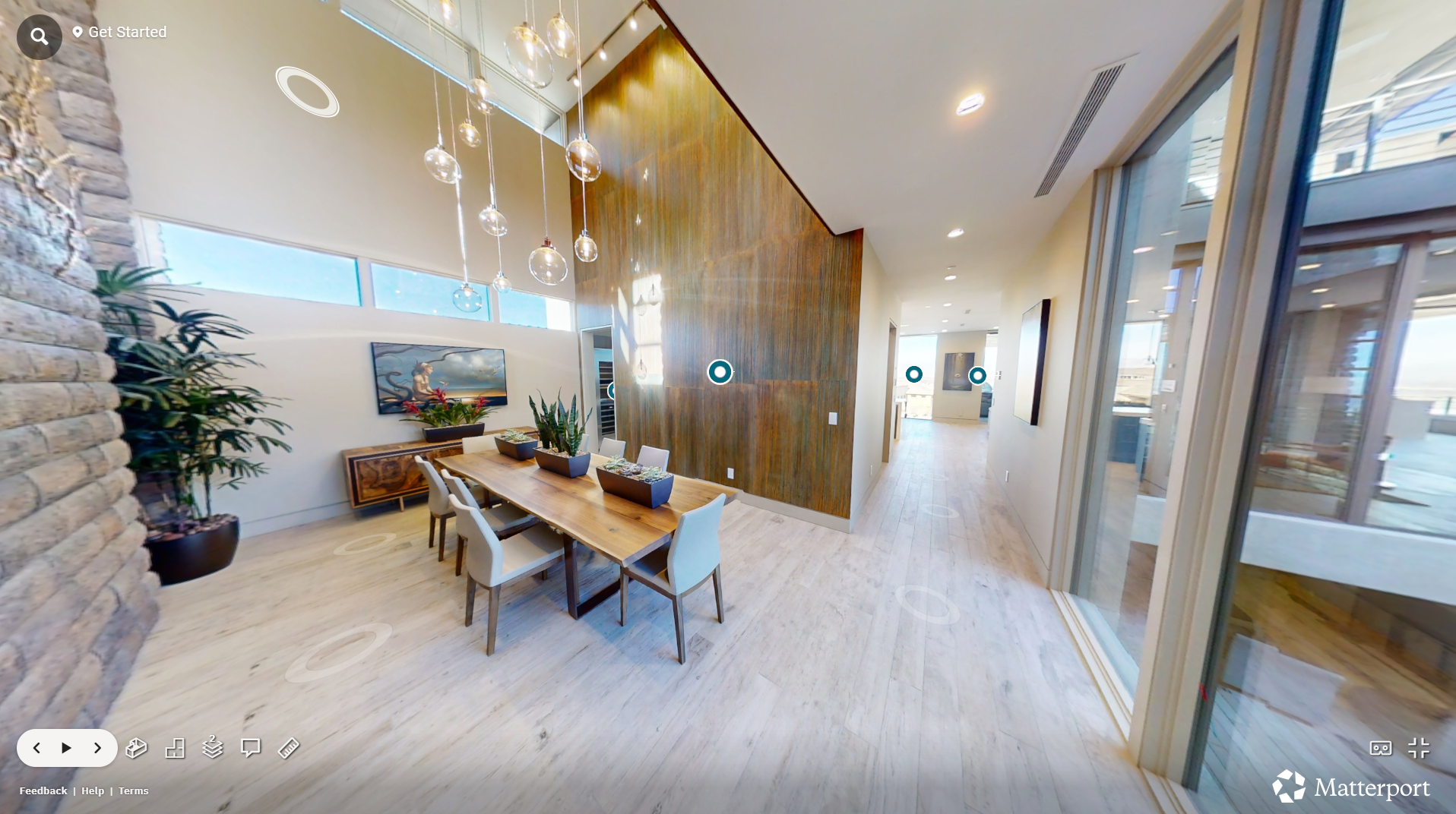 Modern dining area with a wooden table and six chairs, large windows with blinds, a stone wall on the left, potted plants, and a decorative light fixture hanging from the ceiling.