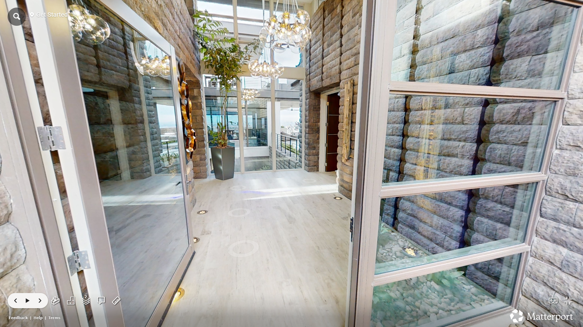 Modern indoor space with stone walls, glass windows, and multiple chandeliers. Green plant in a tall pot near the entrance, and small floor lights.