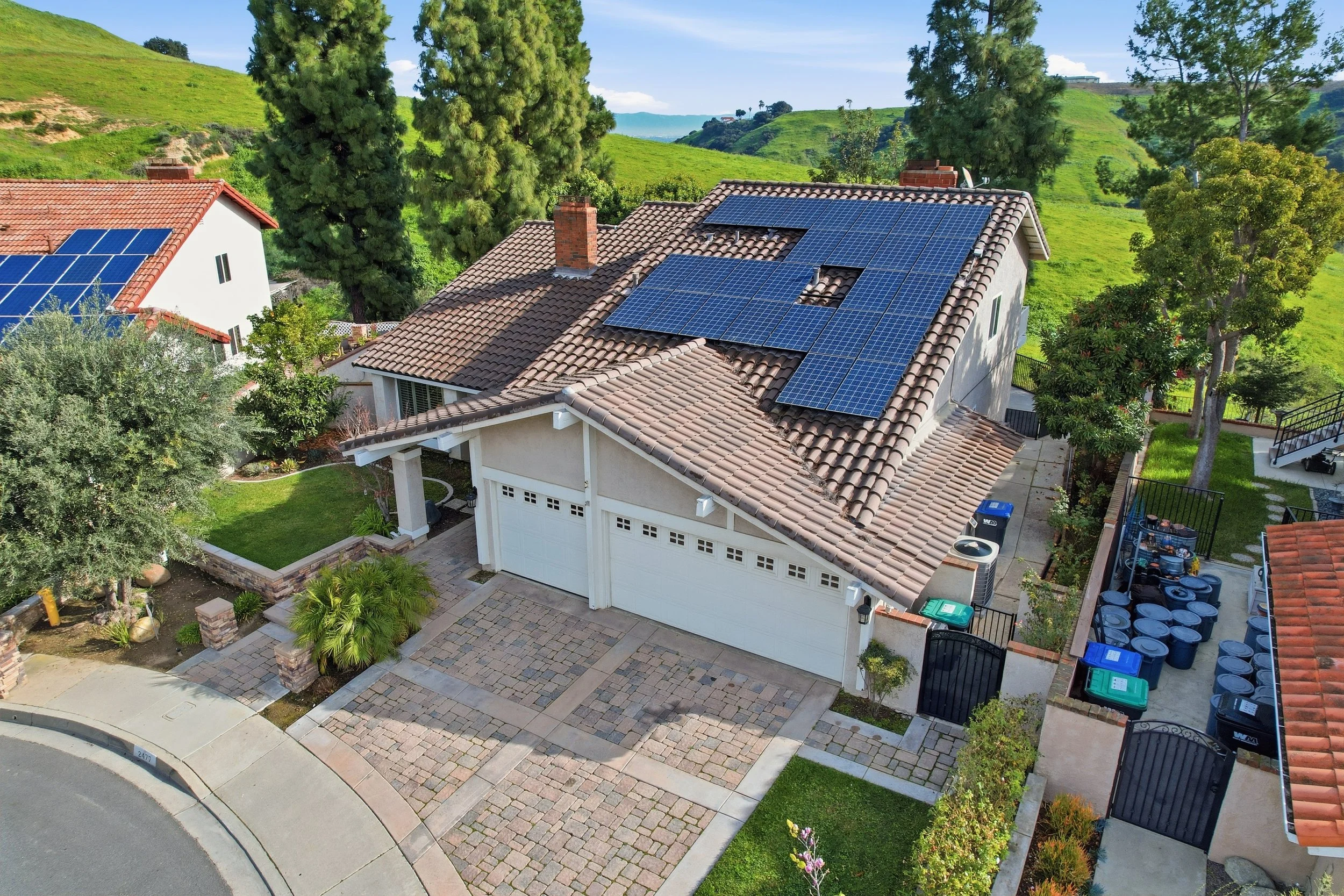 A house with a tiled roof featuring multiple solar panels, a driveway with cobblestone patterns, a small front yard with green grass and plants, and a backyard enclosed by a fence with a viewing deck and several trash bins. The setting is a hilly landscape with trees and green hills in the background.