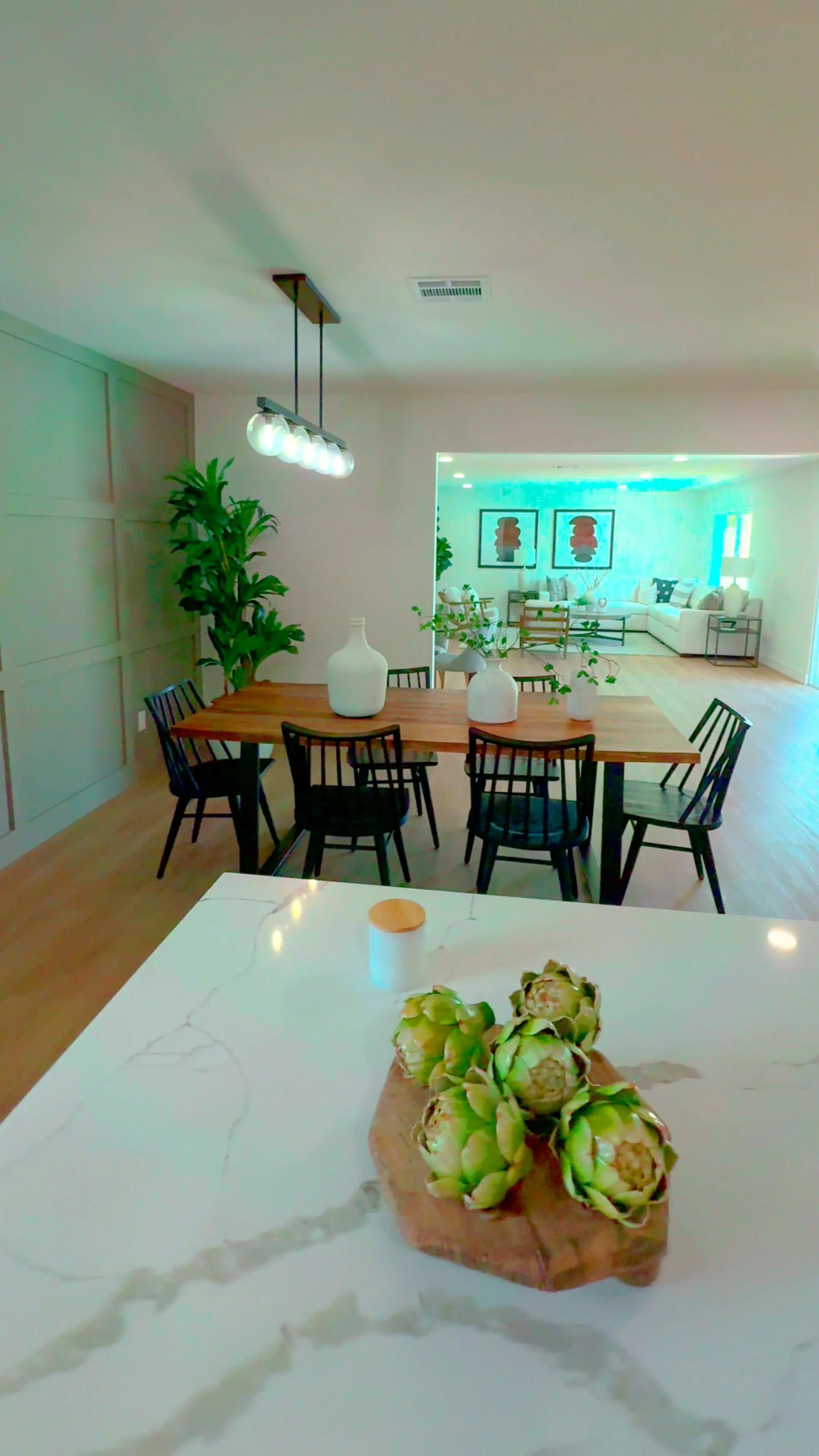 Modern open-plan living and dining area with a marble kitchen island in the foreground, a wooden dining table with black chairs, decorative vases with greenery, and a cozy seating area beyond.
