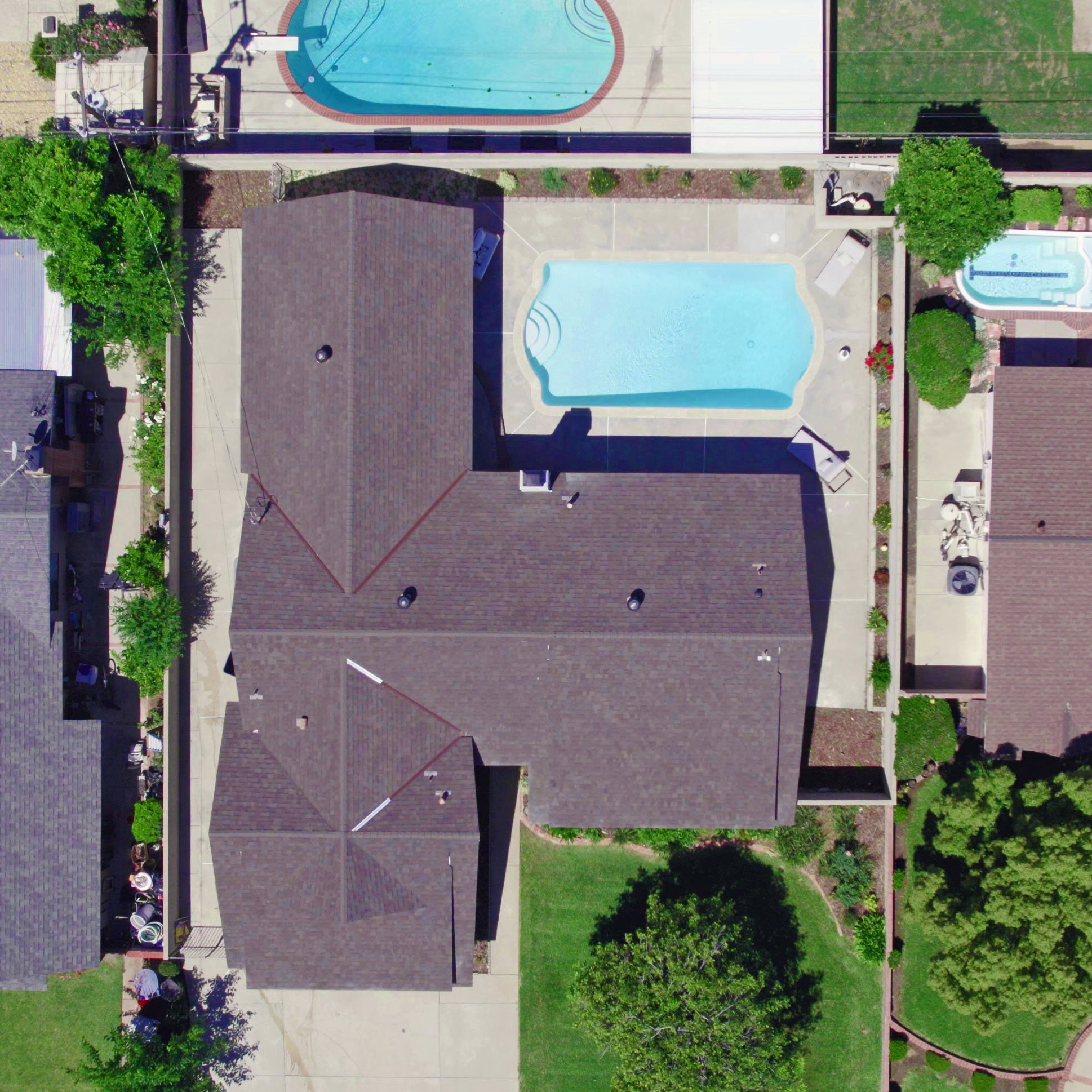 Aerial view of a residential backyard with a swimming pool, a house with a brown roof, and landscaped greenery.
