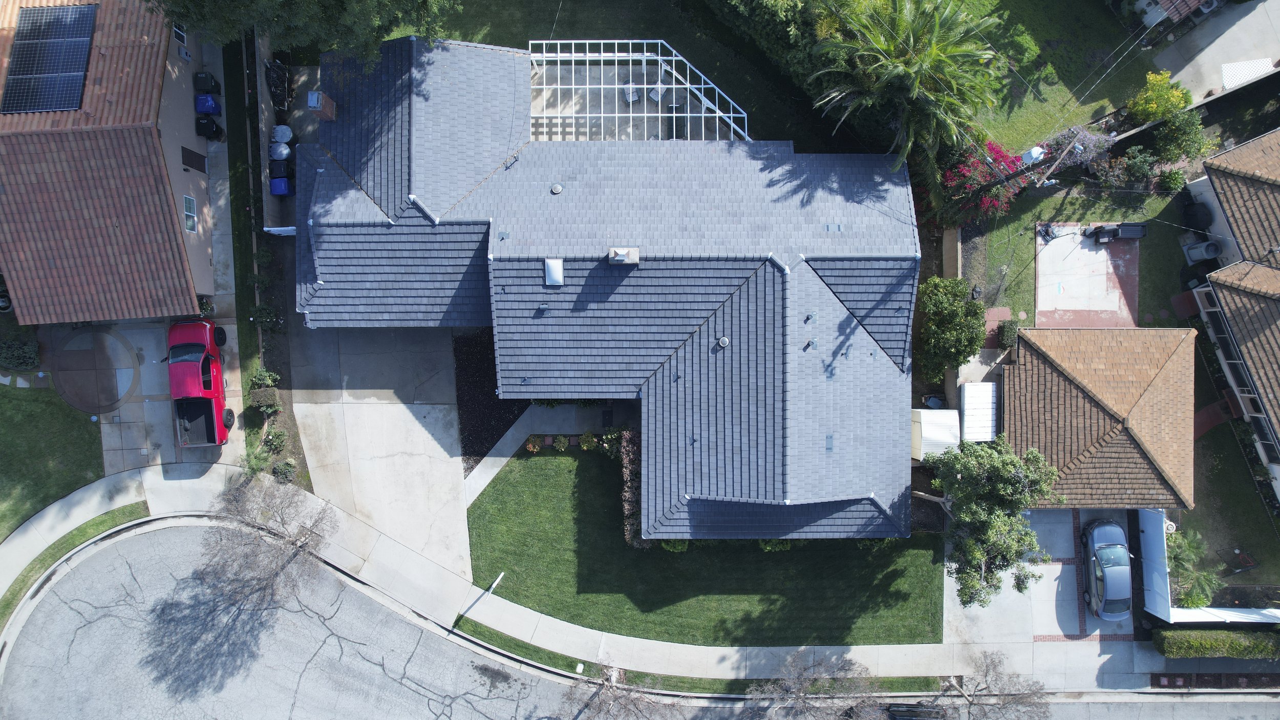 Aerial view of a house with a grey roof, surrounded by a lawn, trees, and neighboring houses with red-tiled and brown roofs. A red truck is parked in the driveway, and there are some palm trees and flowering plants nearby.