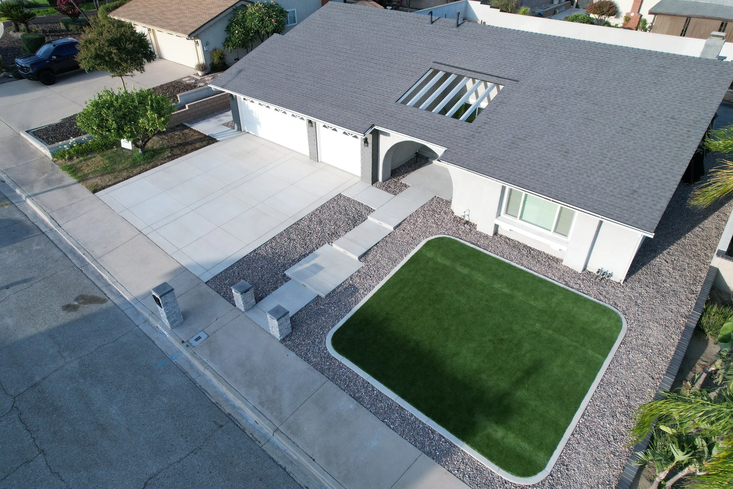 Aerial view of a modern house with a large concrete driveway, a small lawn with green grass, a tree, and a minimalist landscaped yard with gravel and stepping stones.