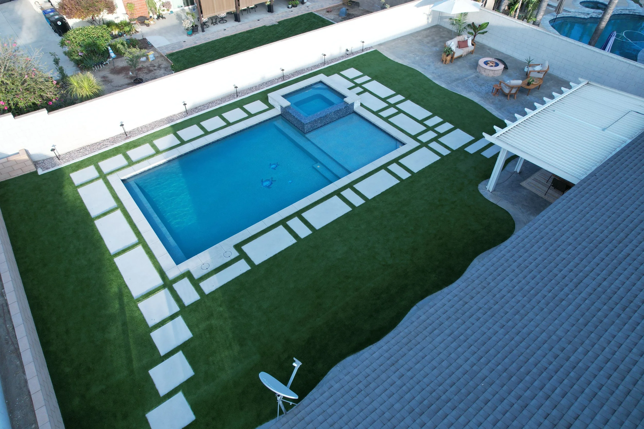 A backyard with a rectangular swimming pool, surrounded by a well-maintained lawn and concrete stepping stones, with a small elevated spa area, outdoor seating, and a satellite dish.