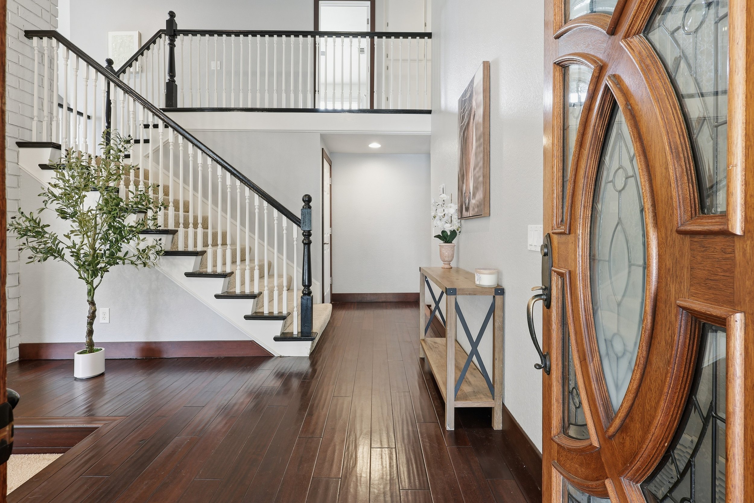 View of a home's foyer with a wooden front door, a console table with decor, a staircase with black and white railing, a potted plant, and a painting on the wall.