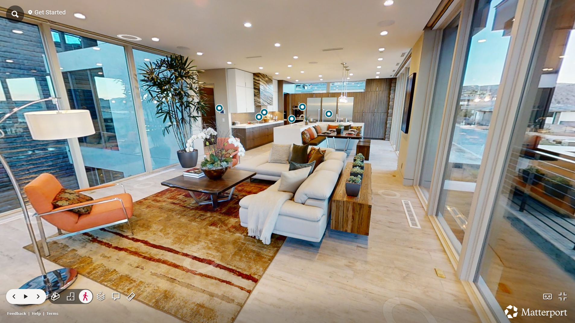 Modern open-concept living room and kitchen area with large windows, white sectional sofa, orange armchair, wooden coffee table, patterned area rug, and various decorative pillows. The kitchen has white cabinets, a kitchen island, pendant lighting, and wooden accents.