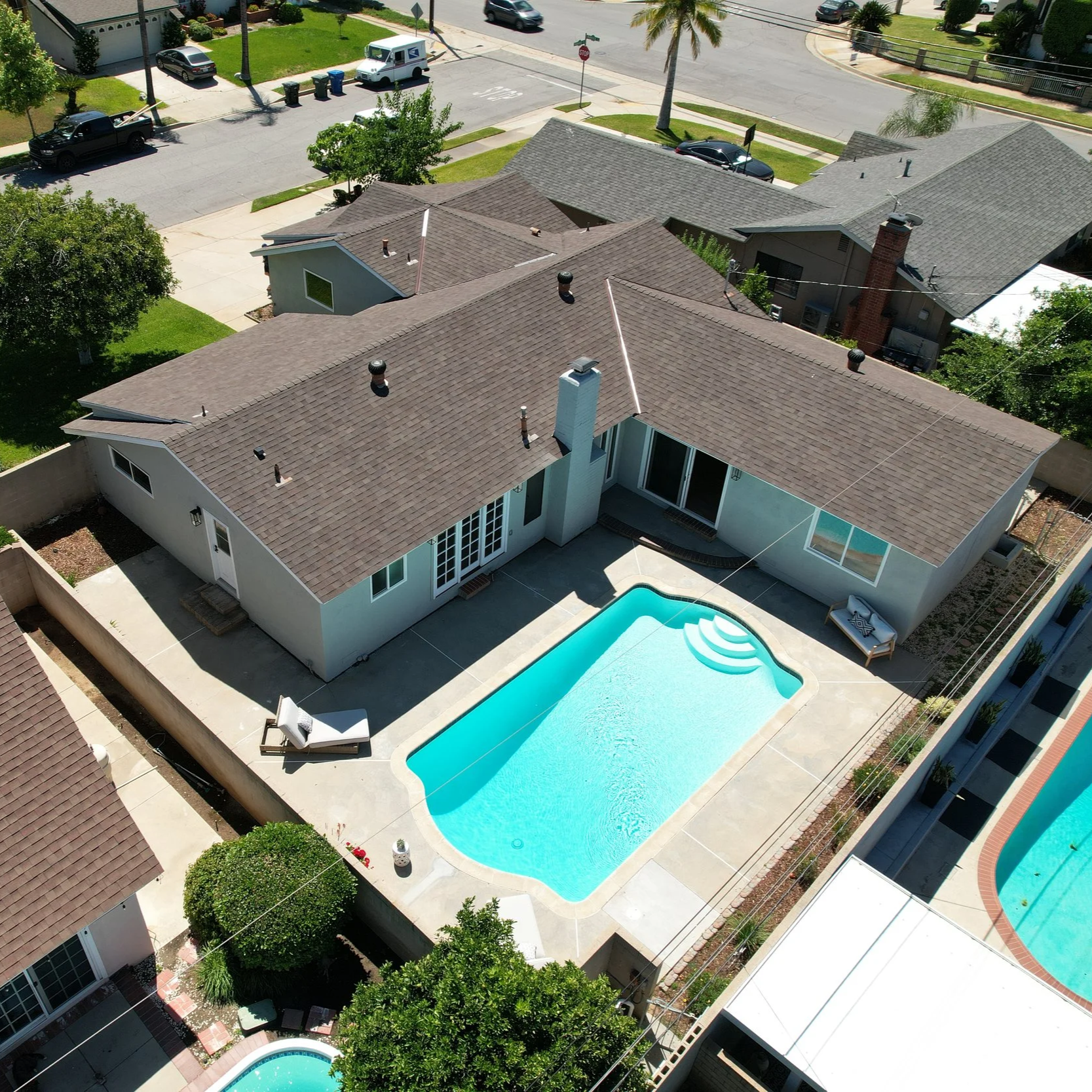 A backyard swimming pool area adjacent to a single-story house with a gray roof, surrounded by concrete and a fence. Several lounge chairs are on the patio, and the pool has curved steps at one end.