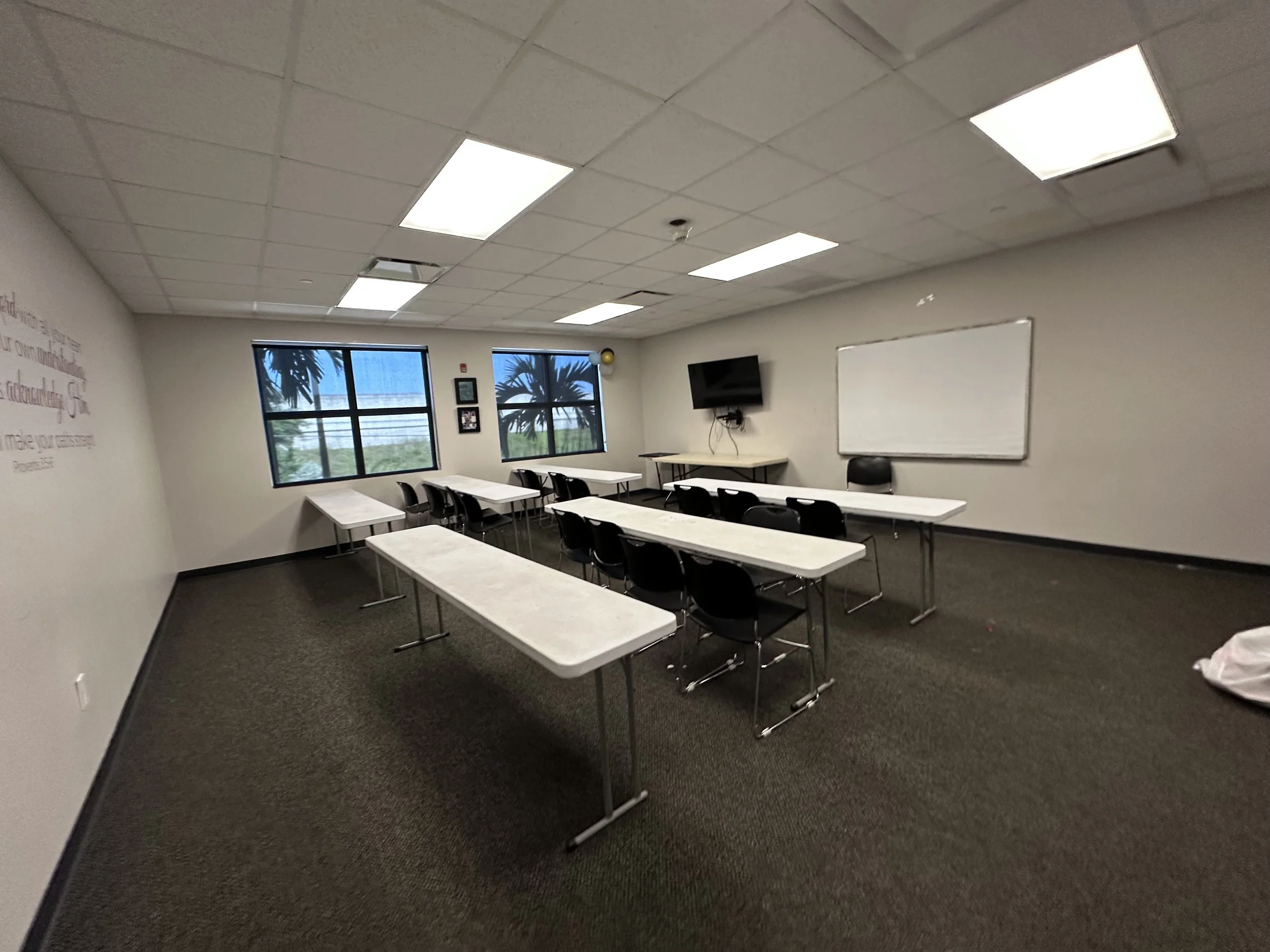 Empty classroom with white tables, black chairs, whiteboard, wall-mounted TV, and windows showing palm trees outside.