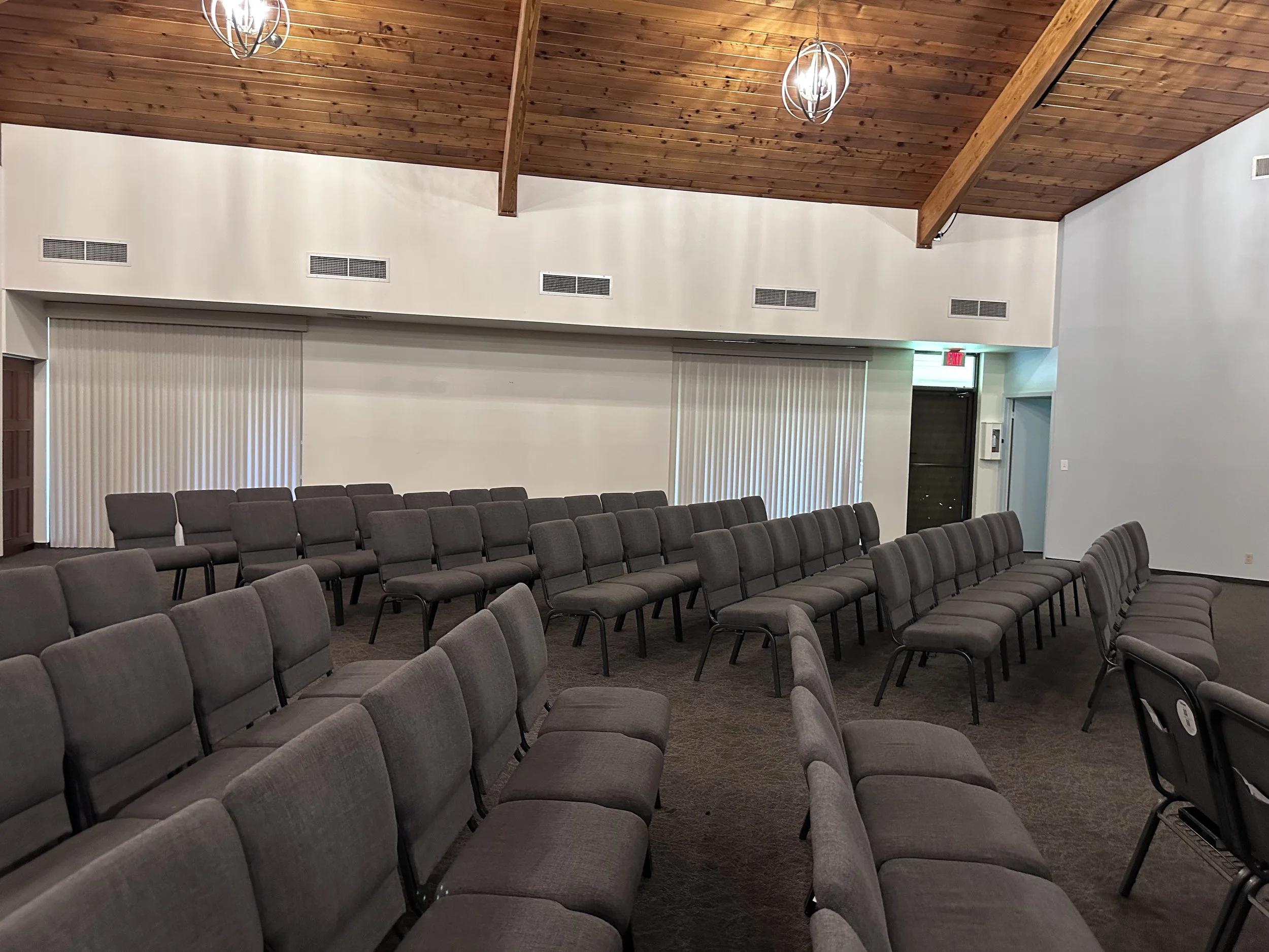 An empty conference or meeting room with multiple rows of gray upholstered chairs arranged on a brown carpeted floor. The room has a vaulted wooden ceiling with two modern light fixtures and a wall of windows covered by white vertical blinds. There are air vents on the upper walls near the ceiling, and an exit door with a green emergency exit sign above it at the back of the room.