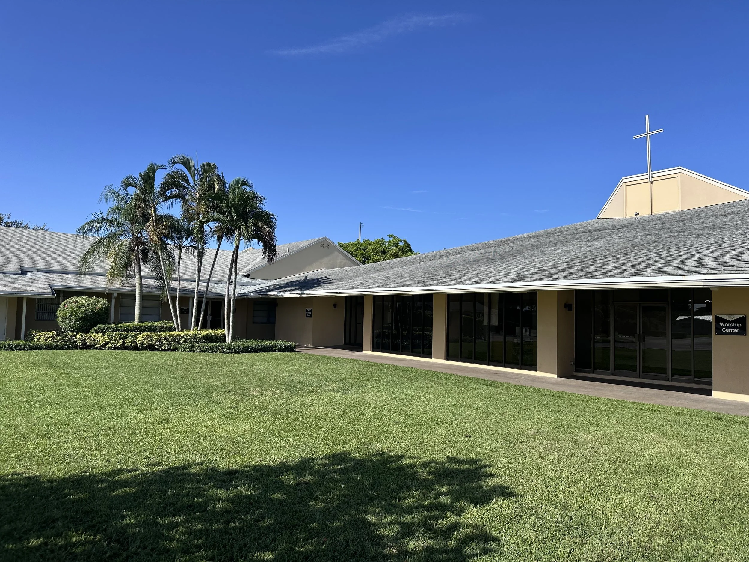 Exterior of a church with a cross on top, large windows, a sign that reads 'Worship Center,' green lawn, palm trees, and a clear blue sky.