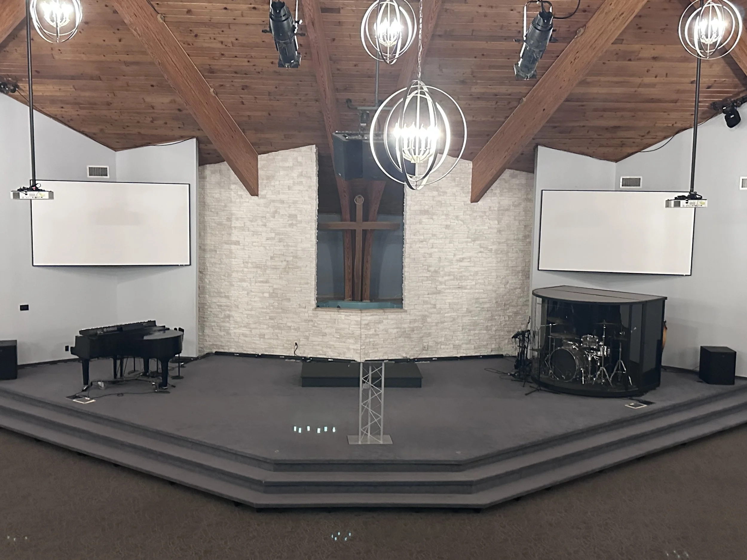 Empty church stage with pianos, drums, large white screens, and hanging chandelier lights, with wooden ceiling and stone wall backdrop.