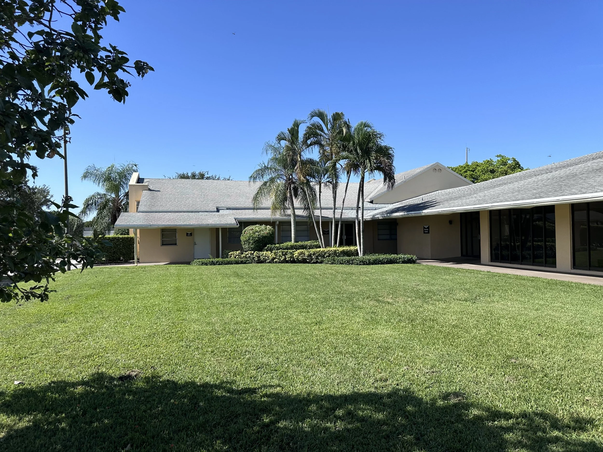 A single-story house with a gray roof, surrounded by a green lawn and tropical trees under a clear blue sky.