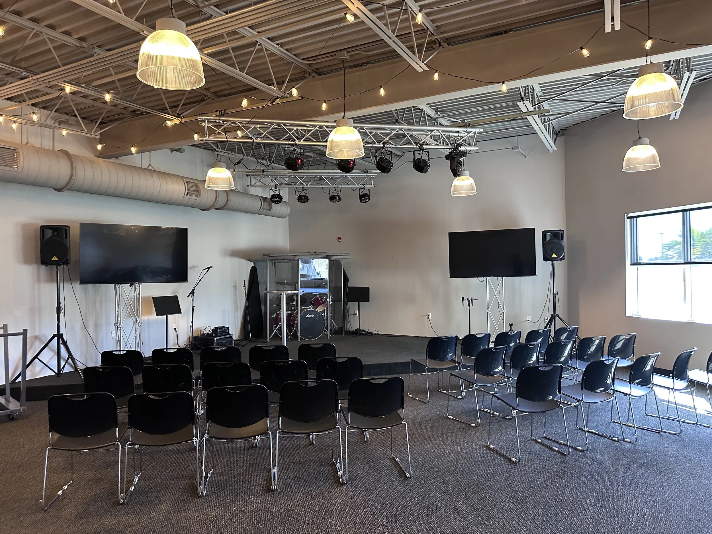 Empty indoor event space with chairs arranged facing a stage. The stage has musical instruments and two large screens on either side, with audio equipment and lighting overhead. There's a window on the right wall, and the ceiling has exposed beams and hanging industrial-style lights.