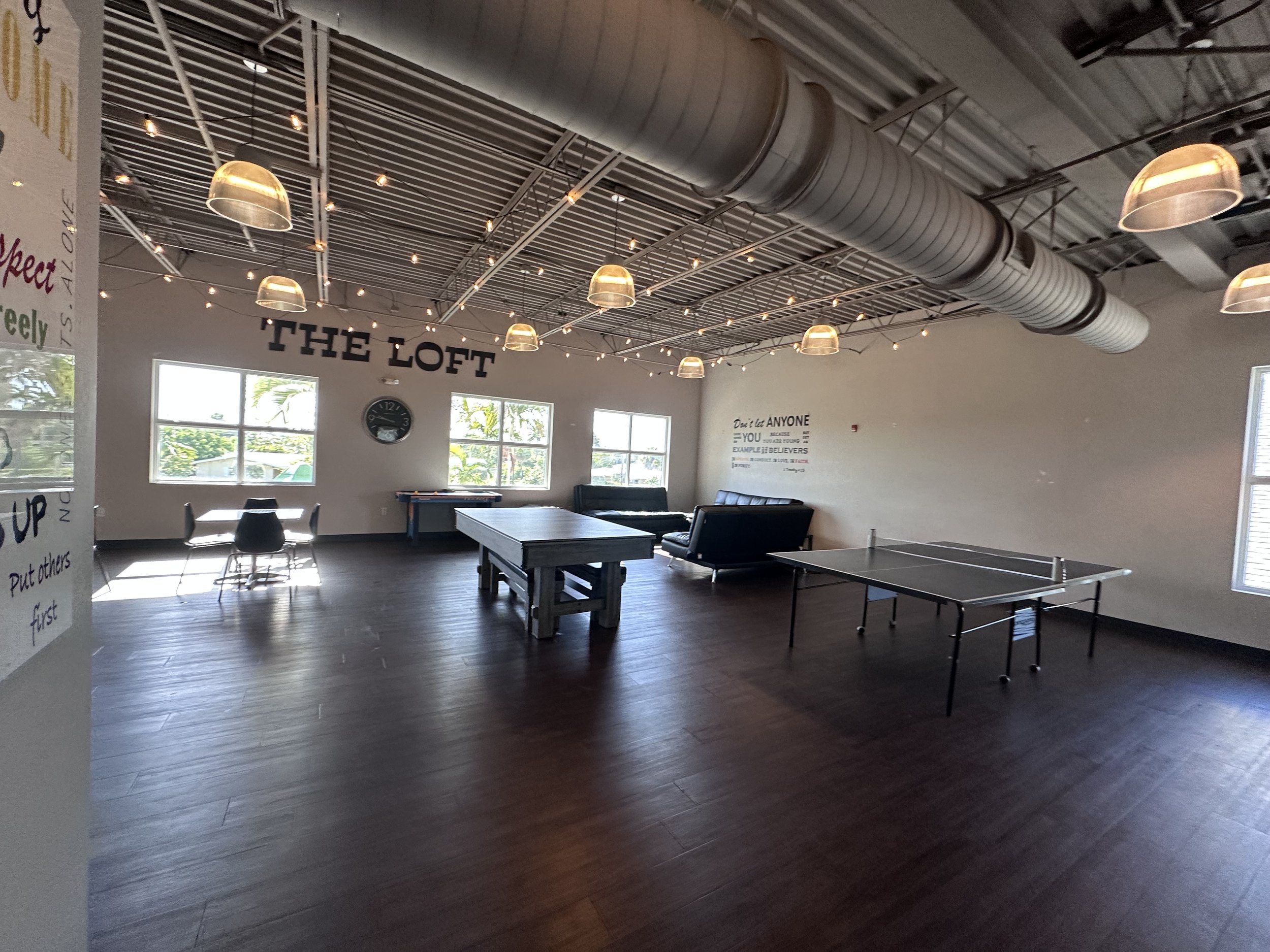 An indoor recreational room with large windows, a ping pong table, a pool table, black couches, and a wall clock. The space has string lights and industrial-style ceiling ductwork, with