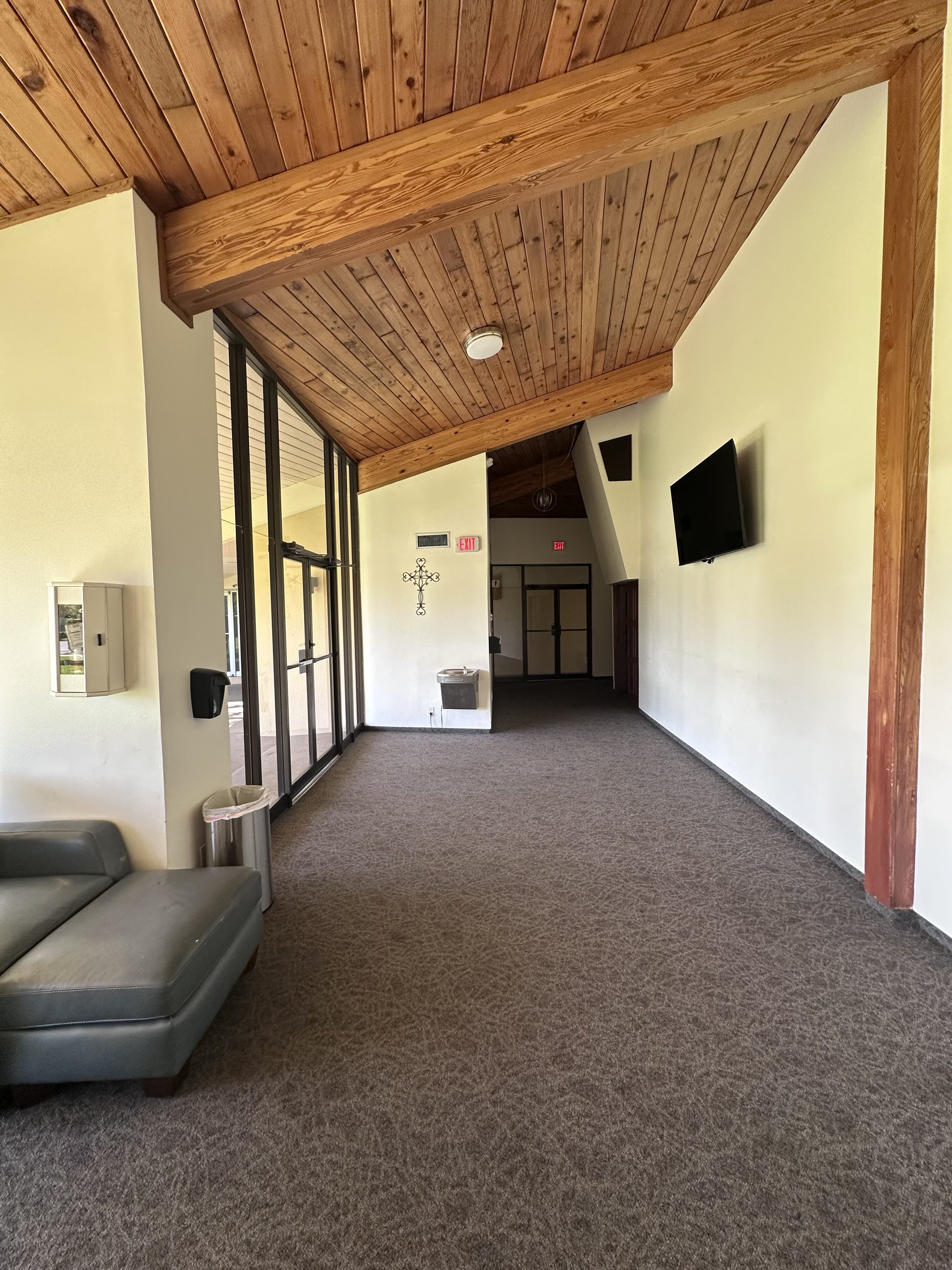Interior of a building with a high wooden ceiling, carpeted floor, mounted TV on a white wall, and glass doors on the left.