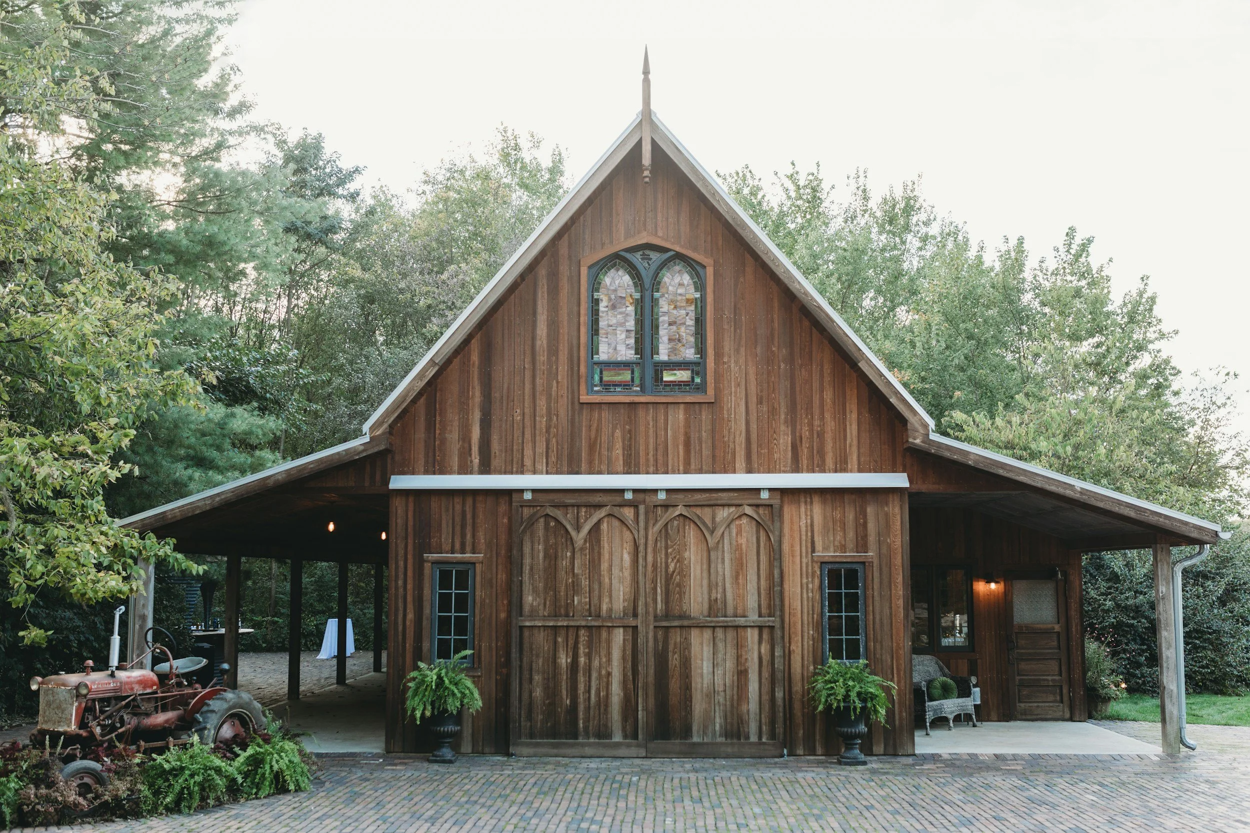 A rustic wooden barn with stained glass windows, surrounded by greenery and trees, featuring a vintage tractor display outside and potted plants near the entrance.