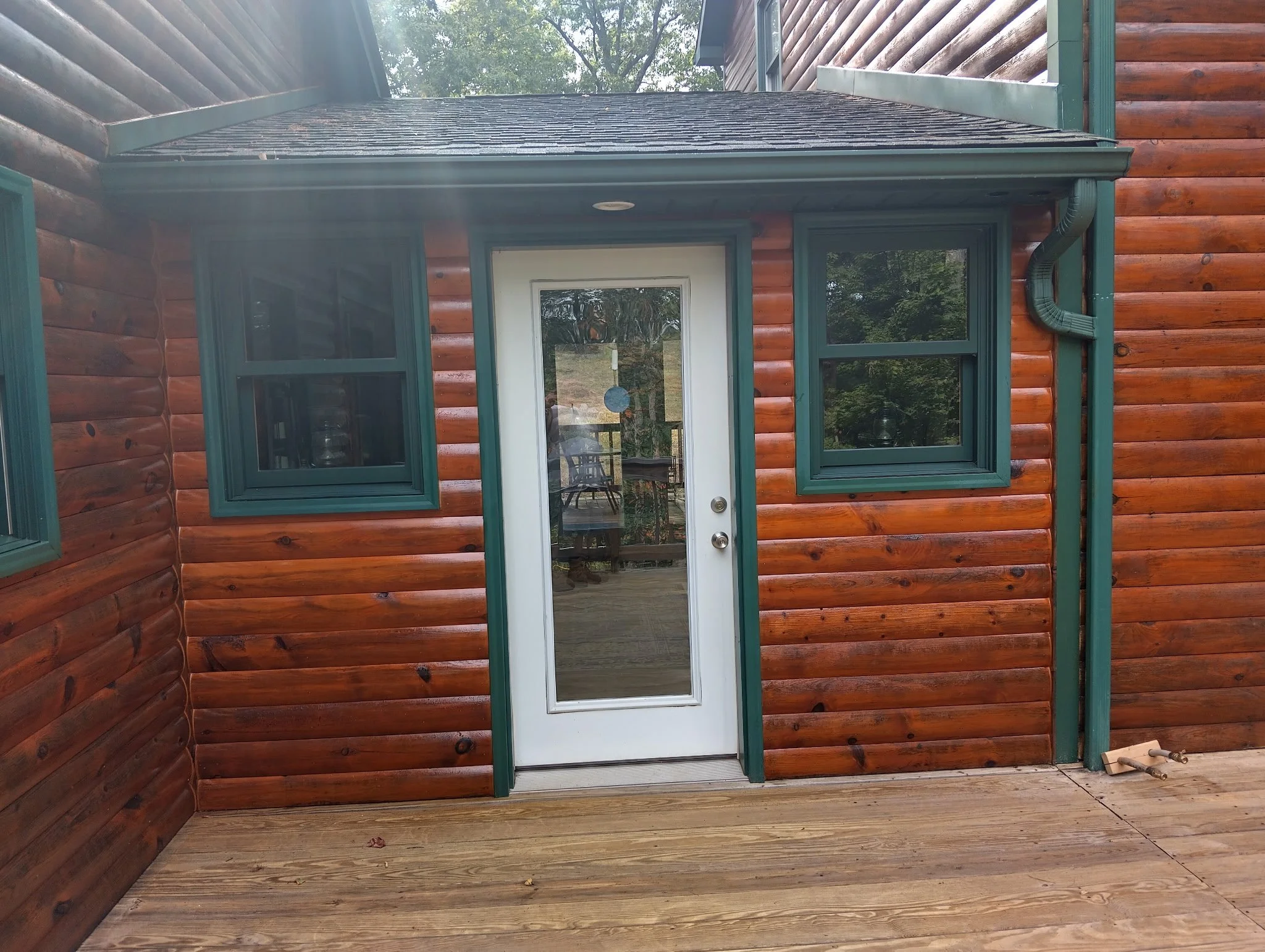 A wooden house with a deck, white door with a glass panel, and two windows with green trim.