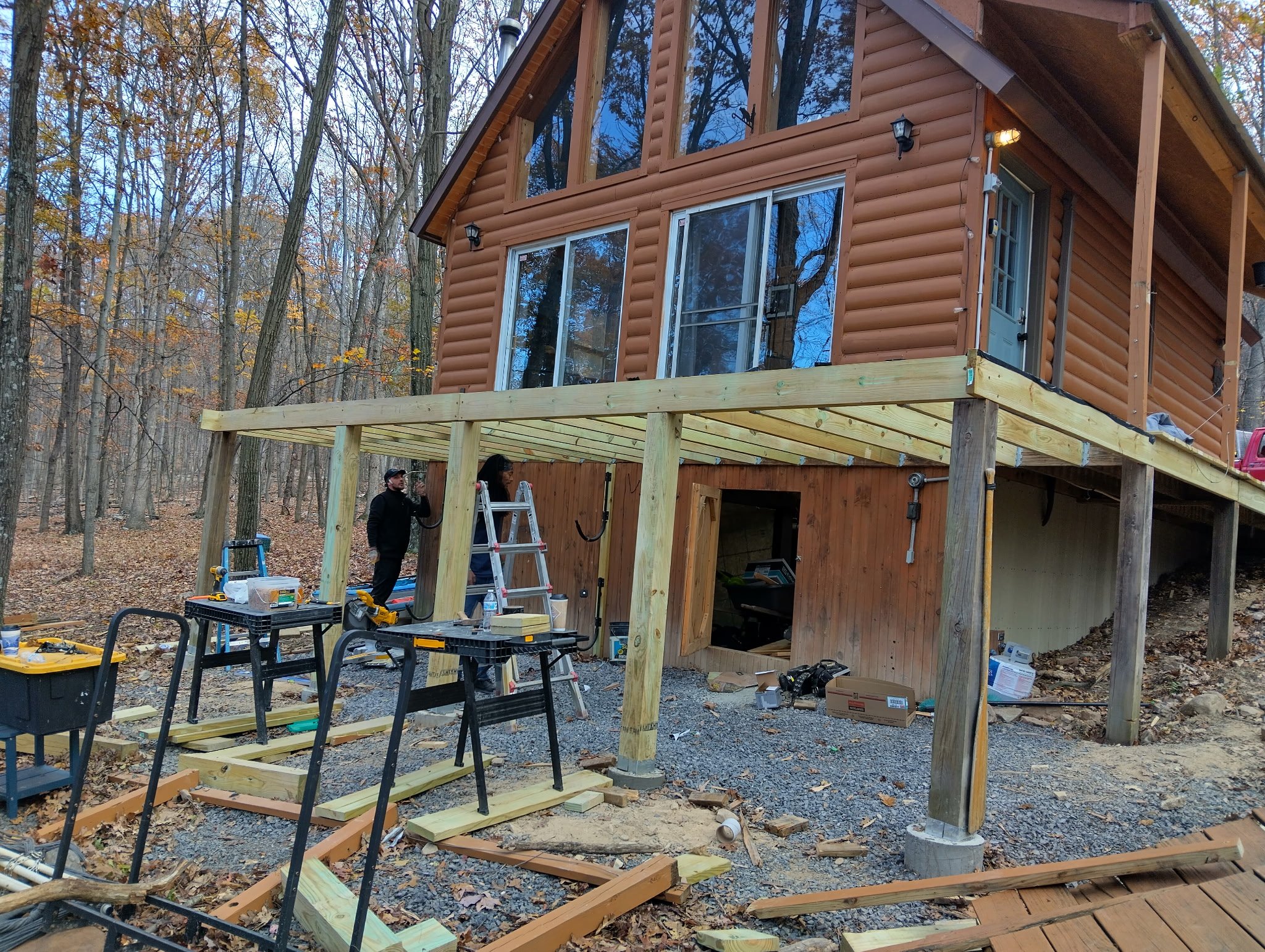 Construction workers installing a wooden deck beneath a high log-style house in a wooded area during fall.