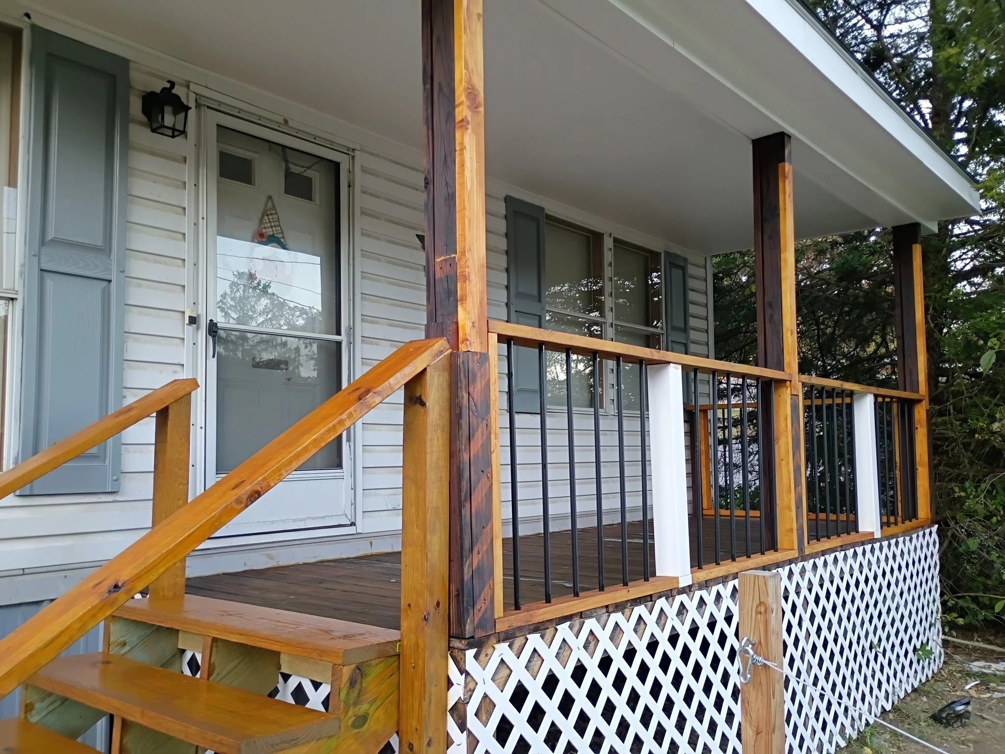 Front view of a house with a small wooden porch, railing, stairs, and lattice skirting. The house has white siding, a screen door, and a window with gray shutters. There are trees and greenery in the background.