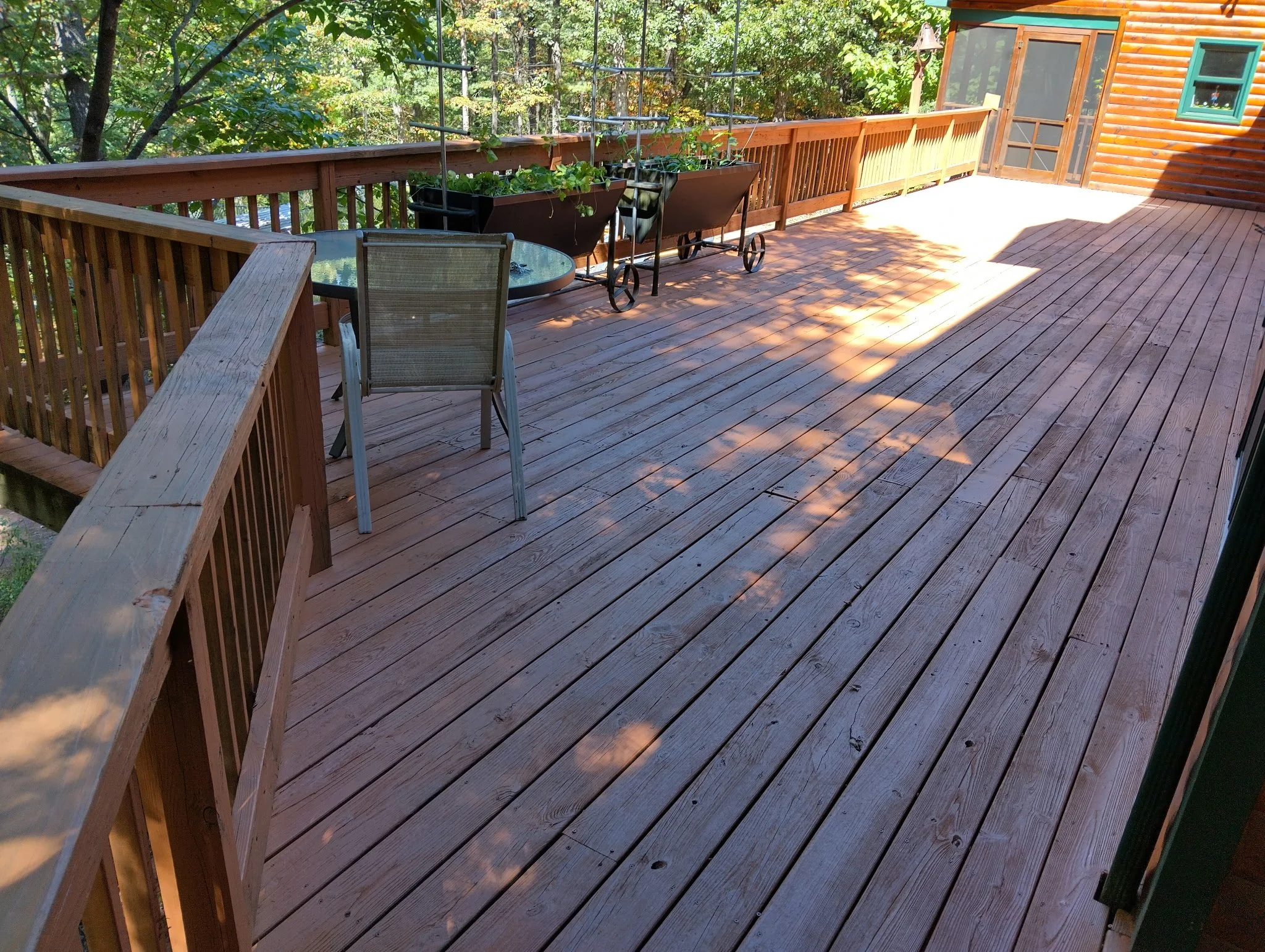 A spacious wooden deck in a forested area, with a small round glass table, a chair, and several potted plants. Sunlight creates shadows on the deck, and there is a screened door leading into a log cabin.