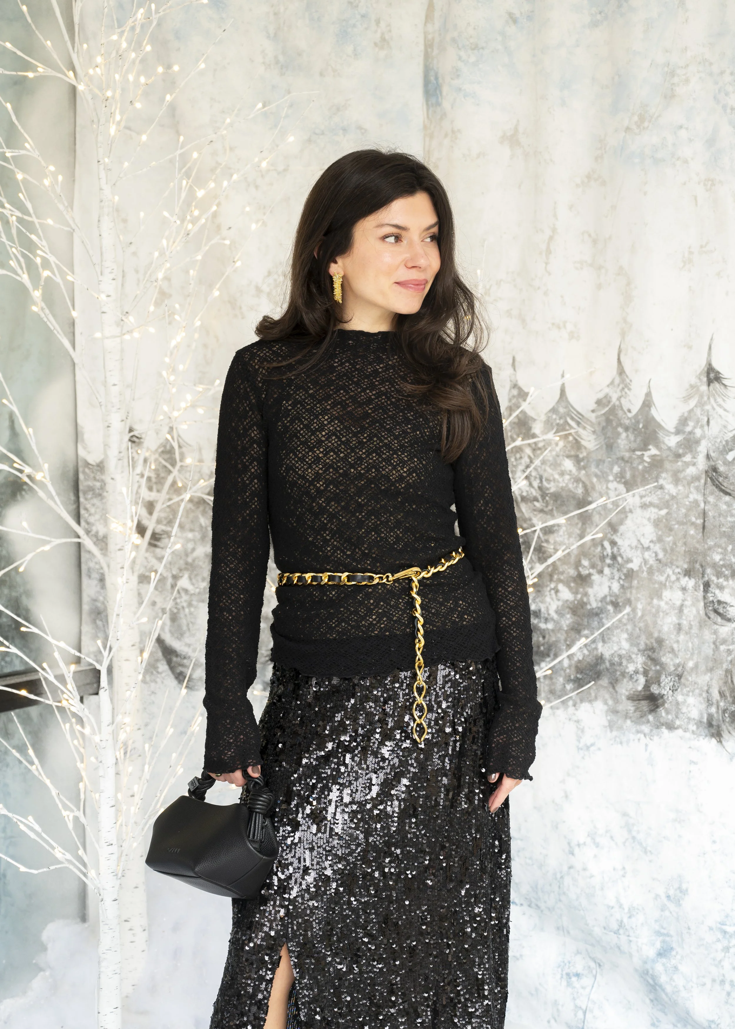 Woman in black lace top and sequin skirt standing in front of winter-themed backdrop with white trees and gray textured wall.