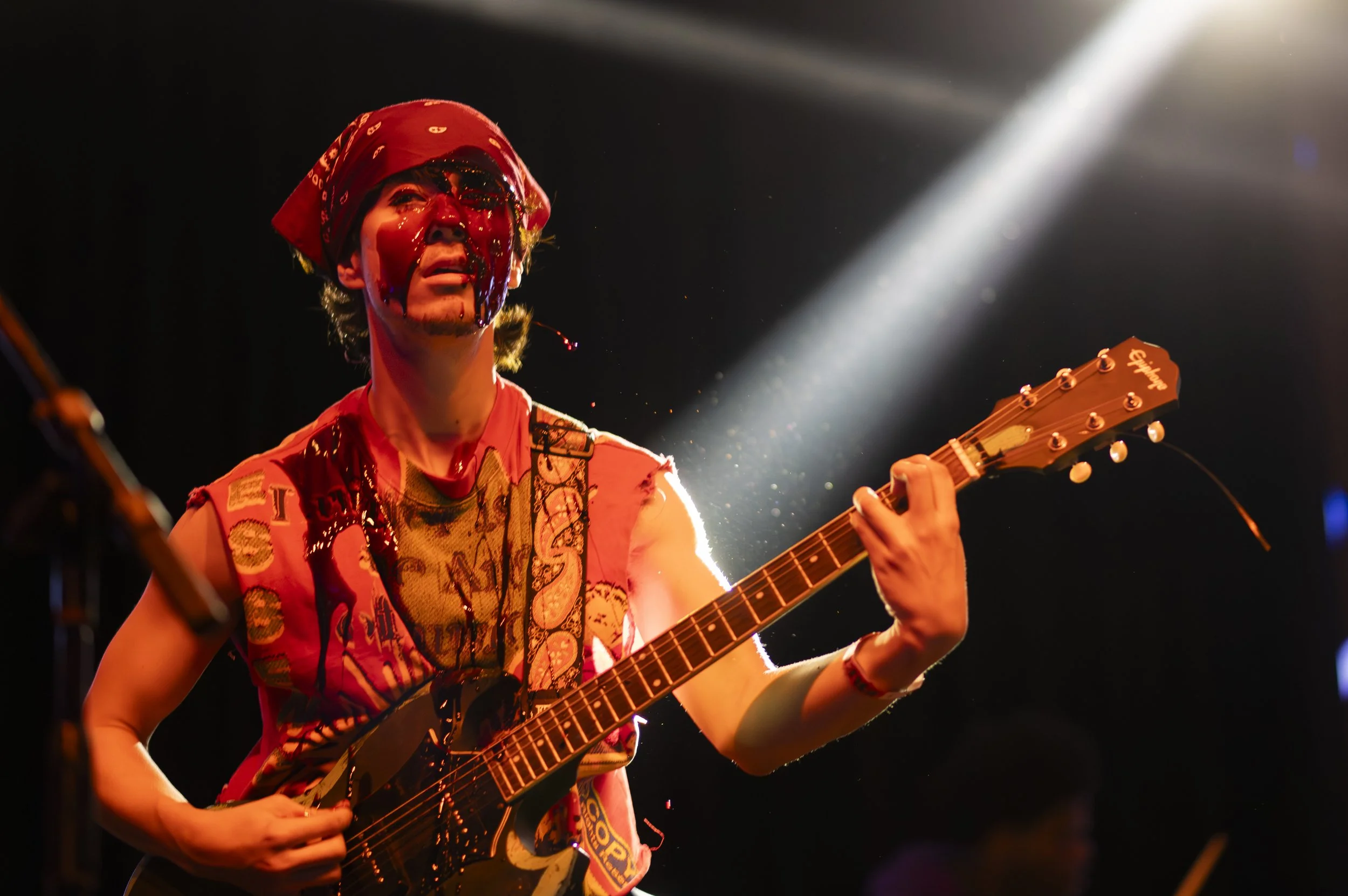 A musician wearing a red bandana and colorful sleeveless shirt playing an electric guitar on stage, with stage lighting illuminating them.