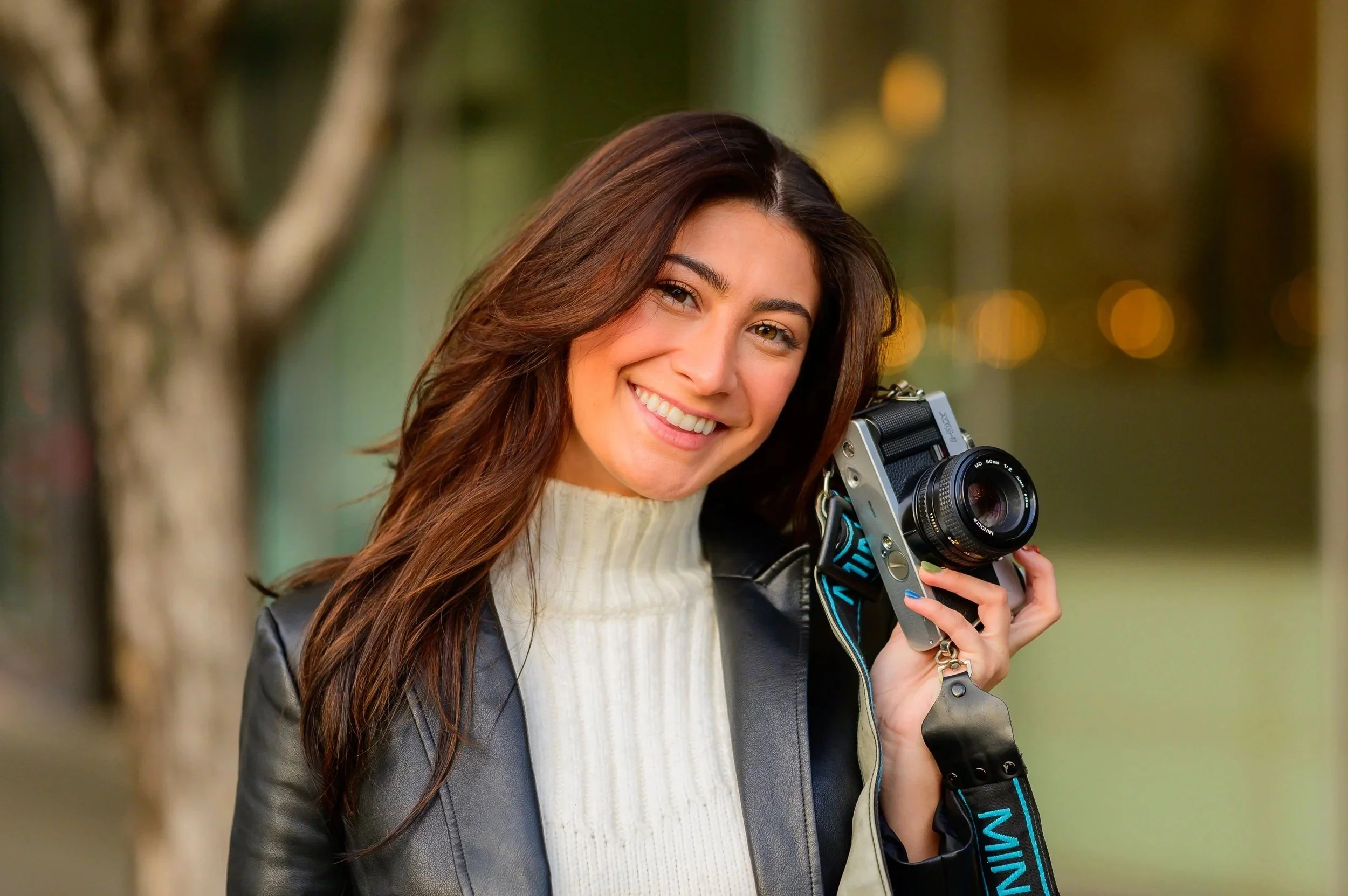 Young woman smiling while holding a camera outdoors with a blurred background of trees and lights.