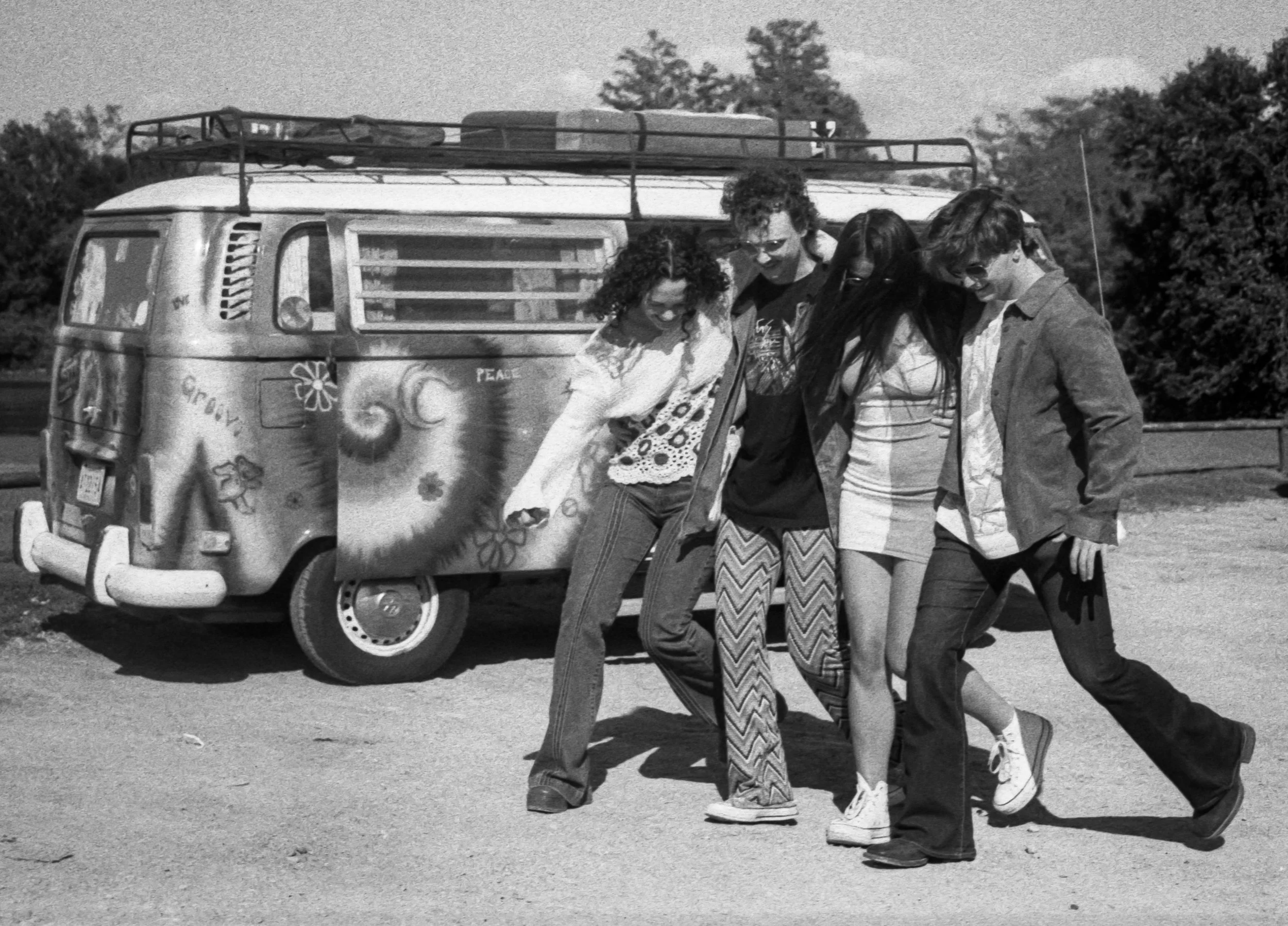 Black and white photo of five young people standing together outdoors next to a decorated Volkswagen camper van. They are smiling and looking down, with their arms linked or resting on each other's shoulders.