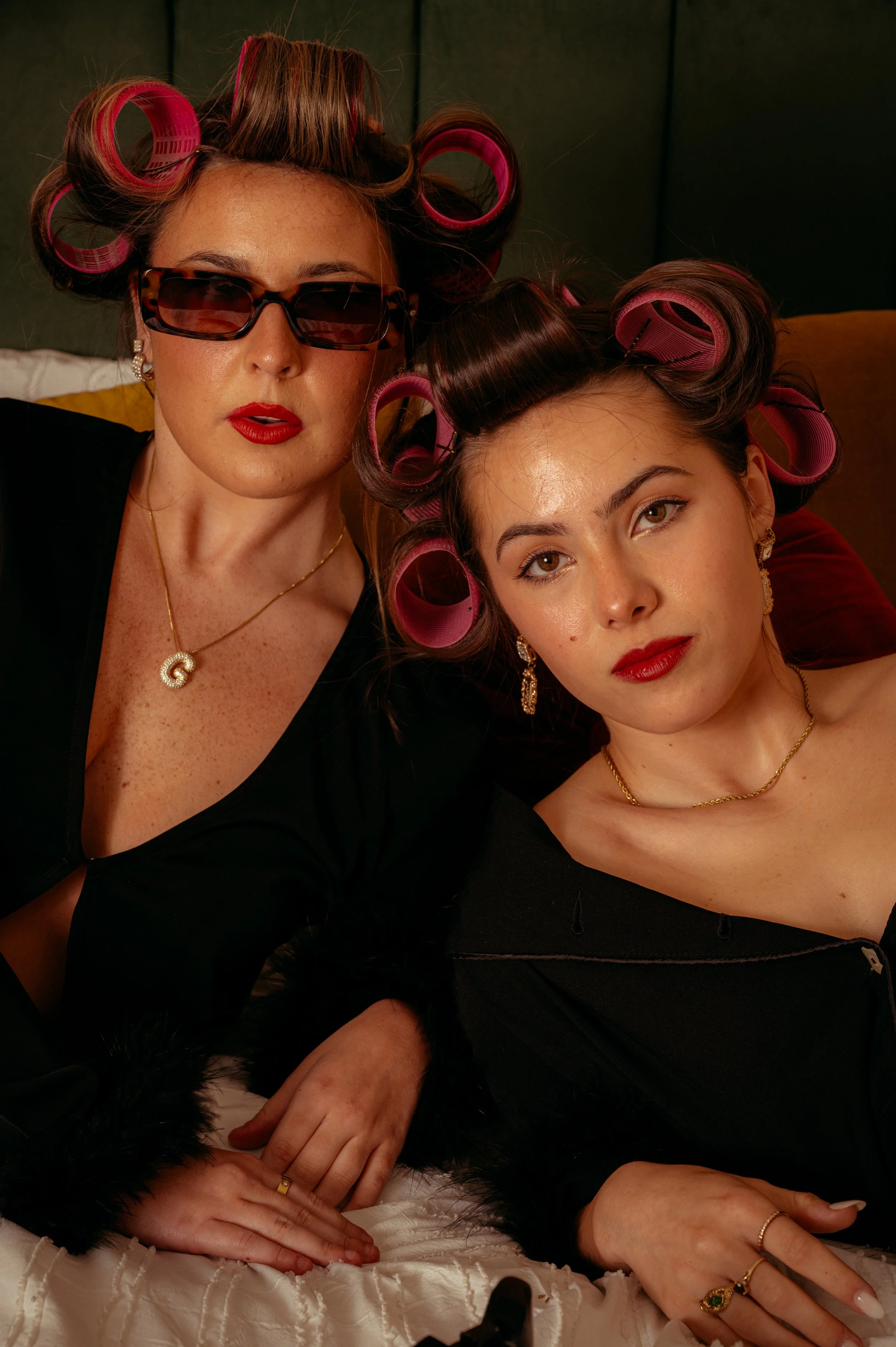 Two women with curlers in their hair, wearing black clothing and jewelry, lying on a bed with a dark background.