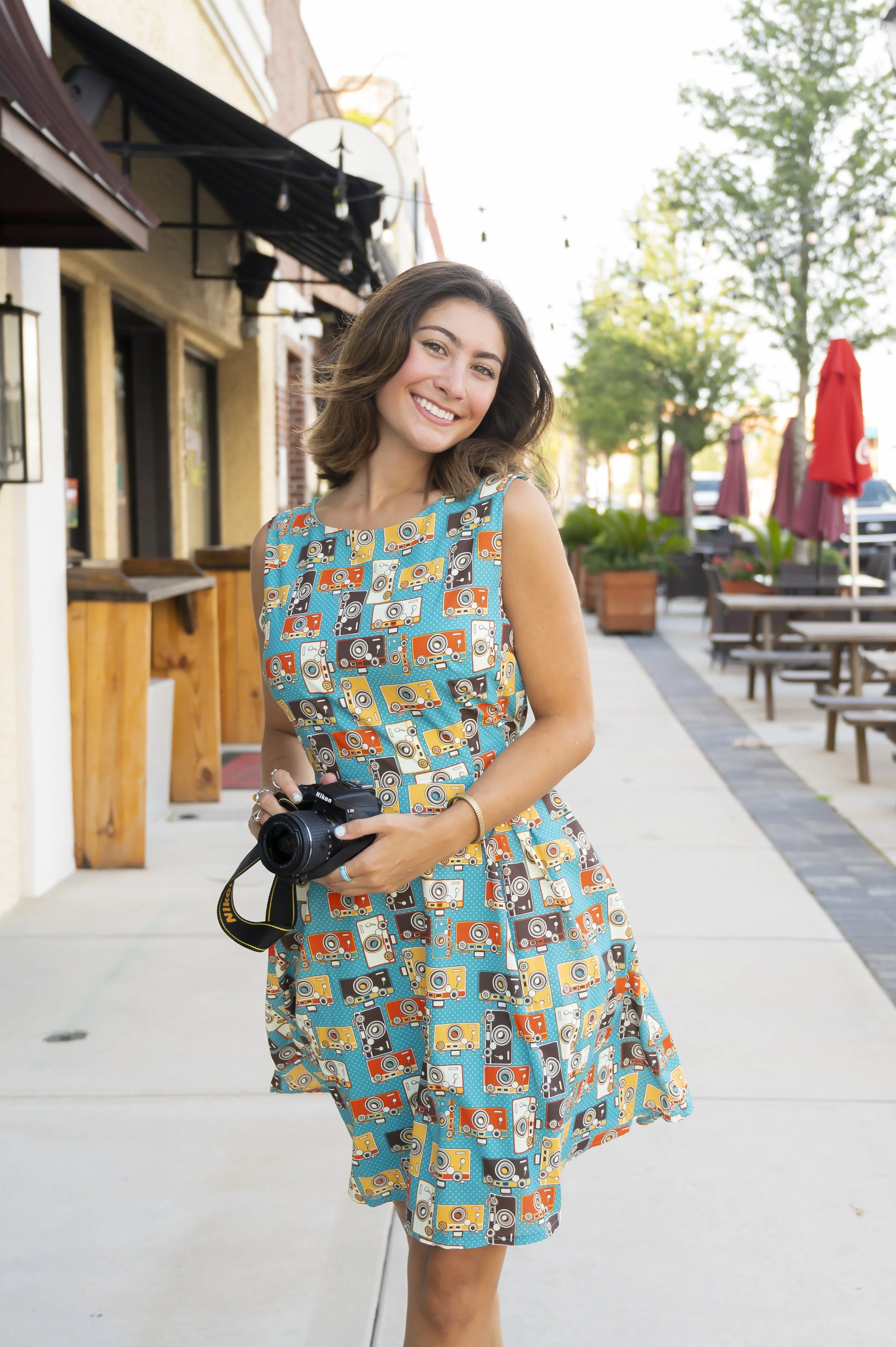 A young woman with shoulder-length brown hair, wearing a colorful sleeveless dress with camera print, holding a DSLR camera, smiling outdoors on sidewalk with patio seating and trees.