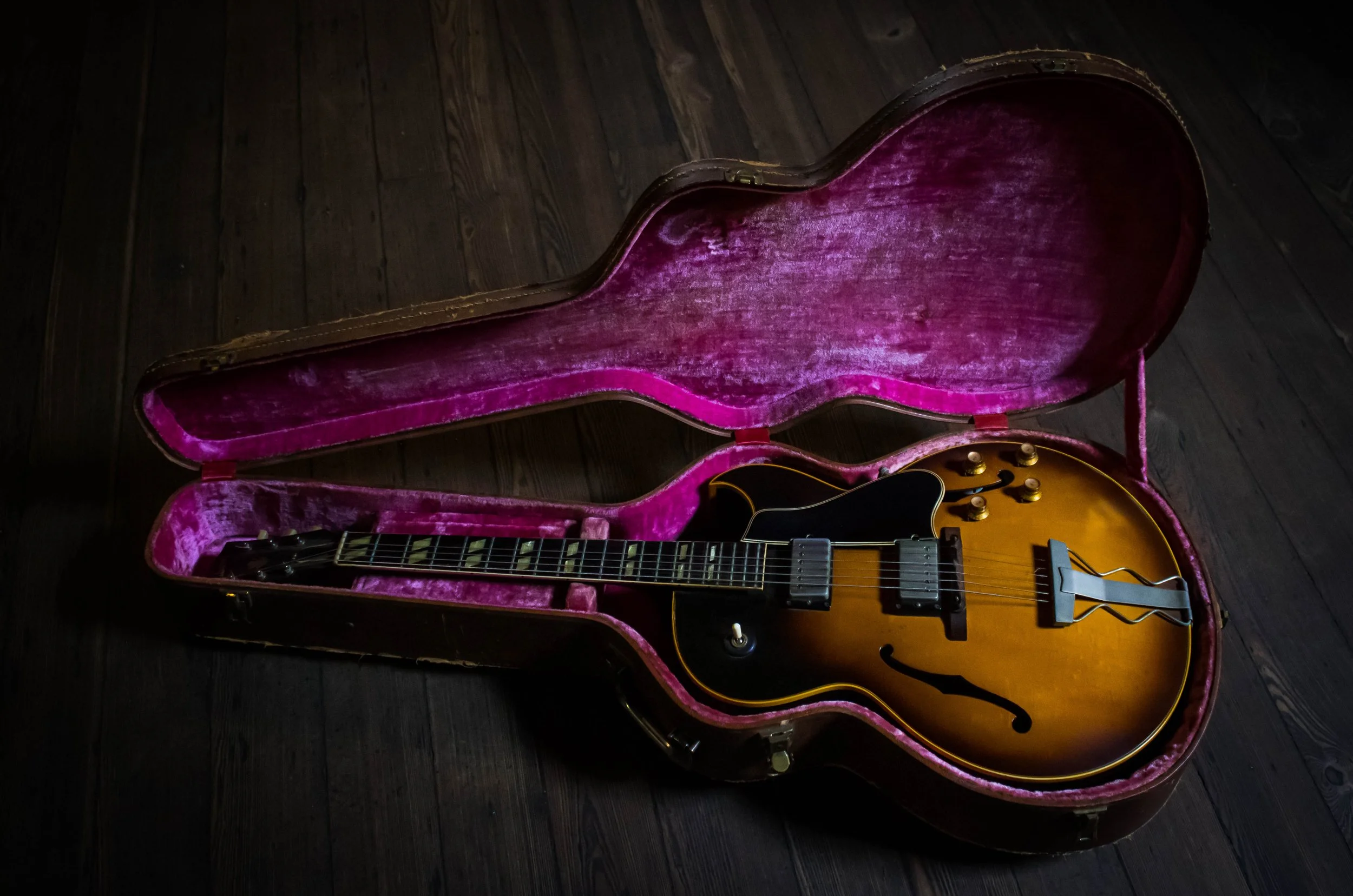 A vintage hollow body electric guitar with a sunburst finish, inside an open pink velvet-lined guitar case on a dark wooden floor.