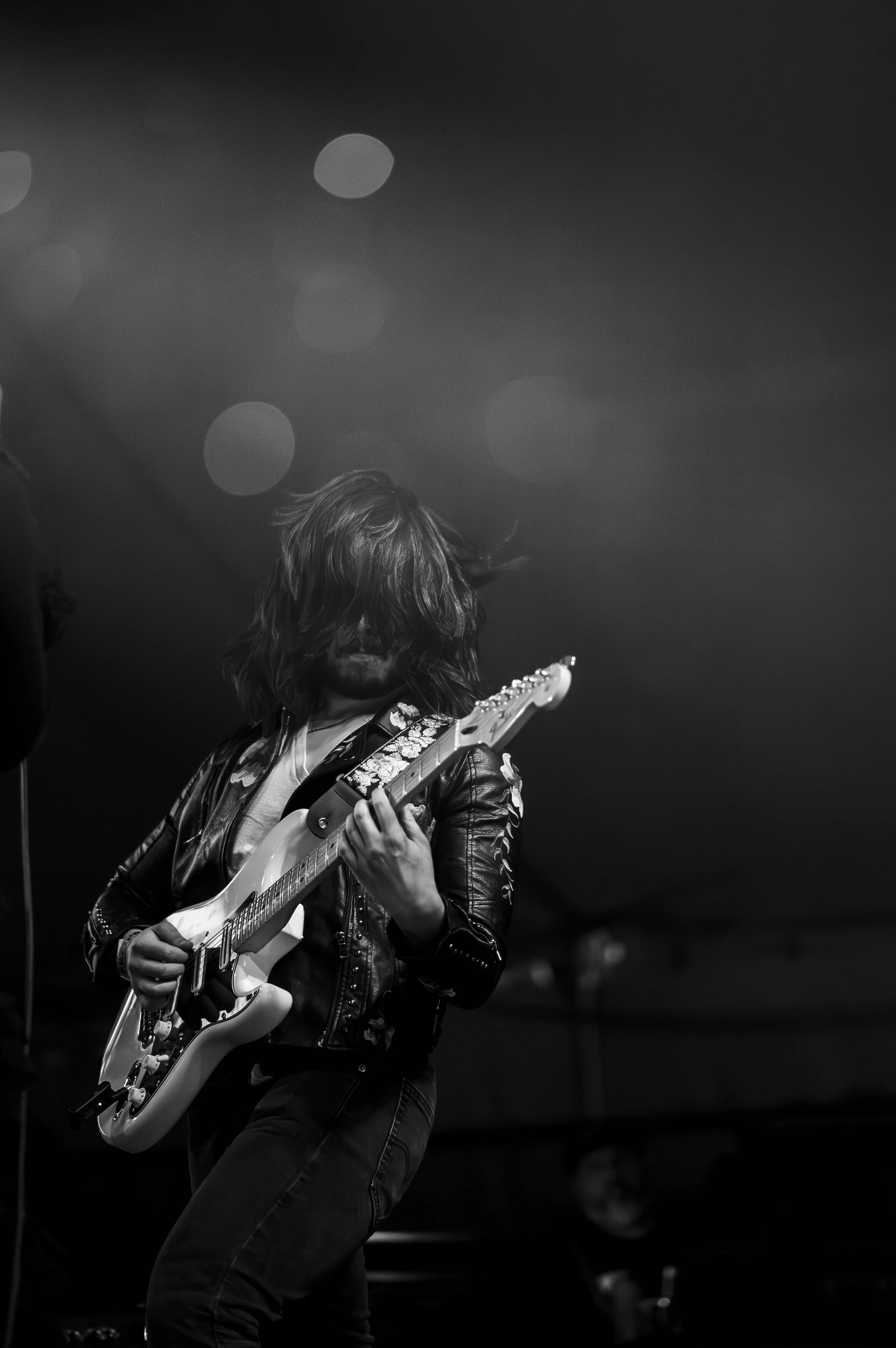 A person with long hair playing an electric guitar on stage in a black leather jacket, with a dark background and soft stage lighting.