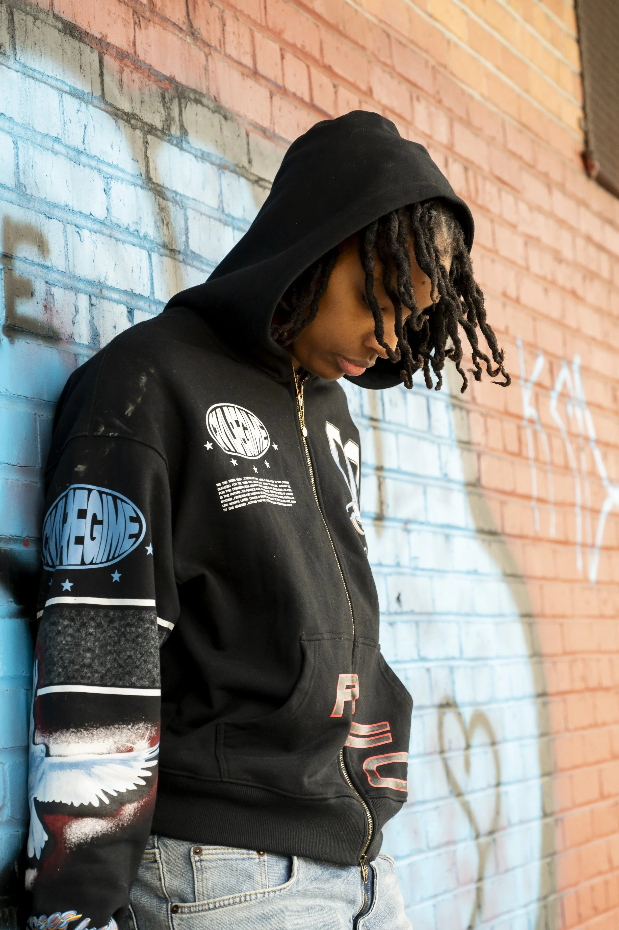 A young person with dreadlocks wearing a black hoodie with patches and graffiti artwork, standing against a colorful brick wall with graffiti tags.