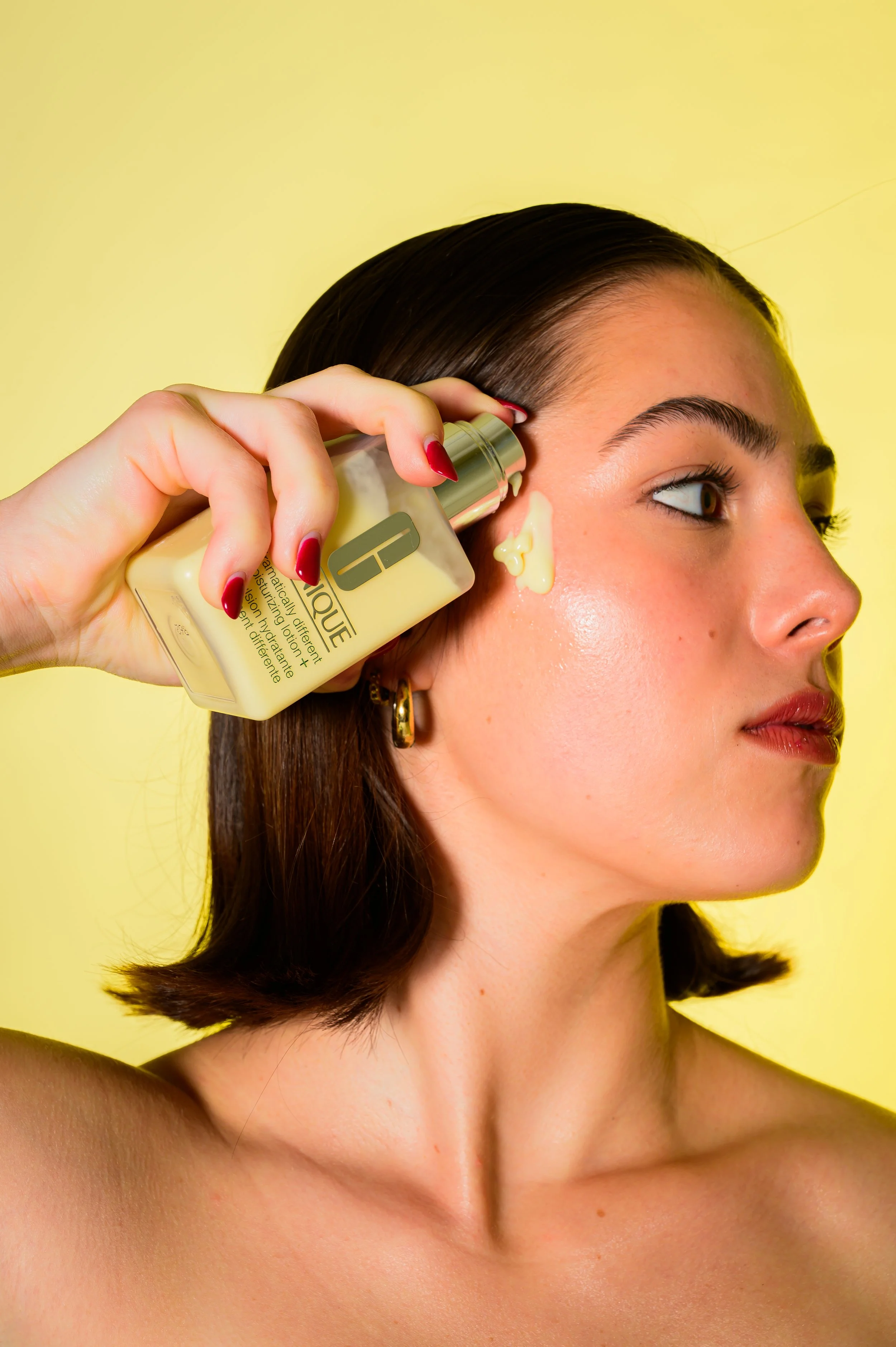 A woman with short dark hair applying a yellow cosmetic product to her face against a yellow background.