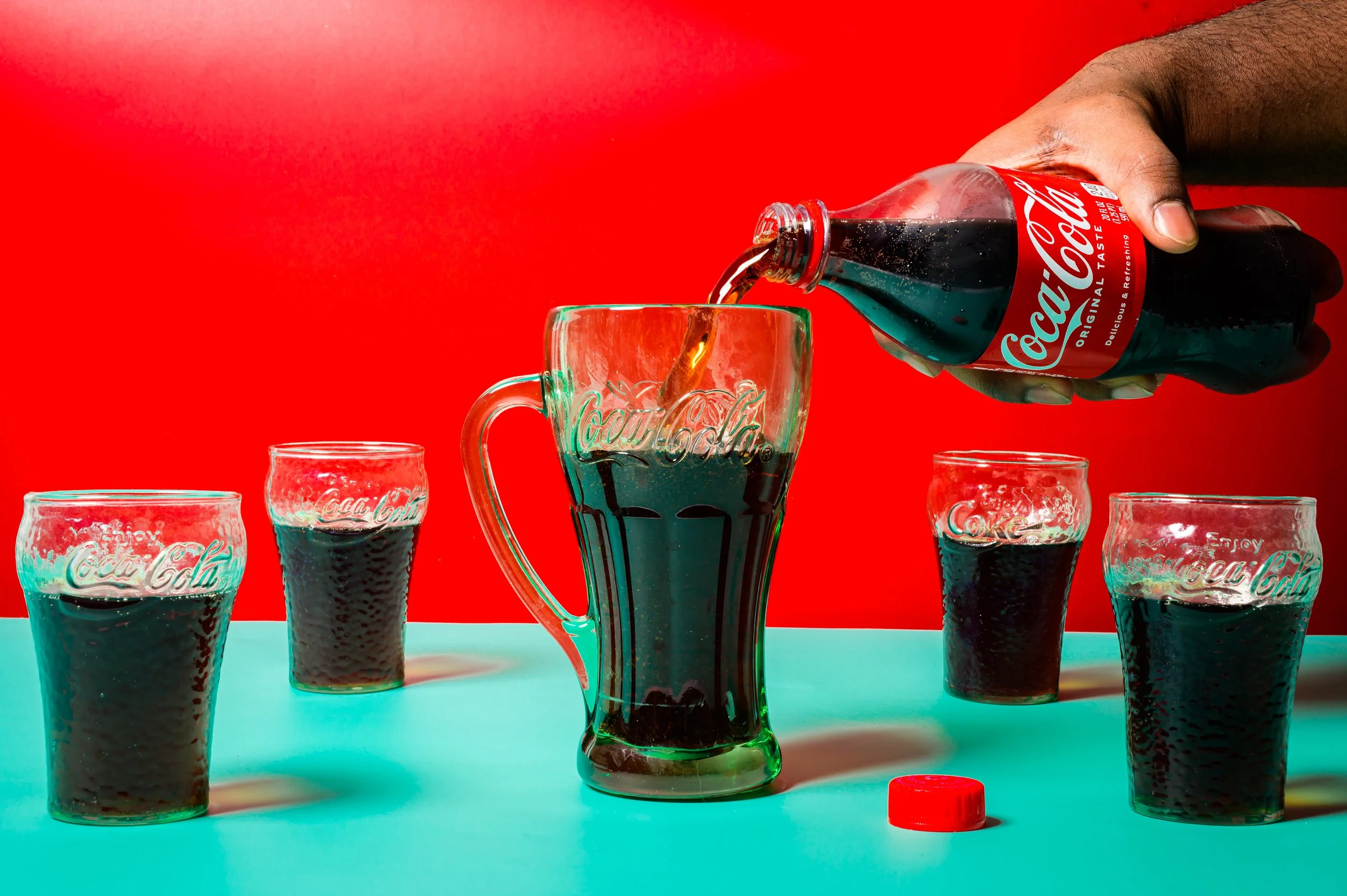 A hand pouring Coca-Cola from a glass bottle into a glass mug with the Coca-Cola logo, surrounded by four smaller Coca-Cola glasses on a turquoise surface with a red background.