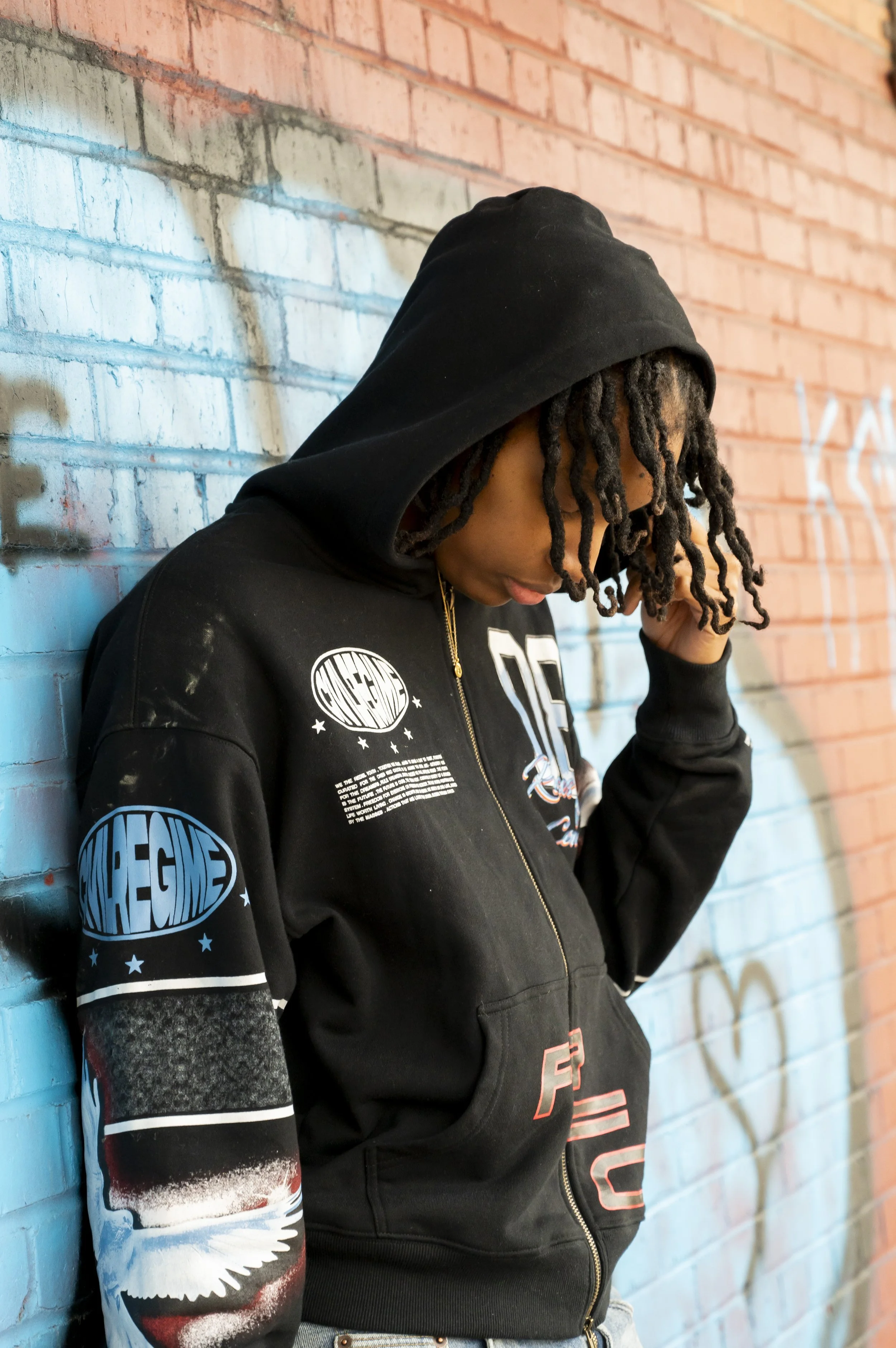 A young person with dreadlocks wearing a black hoodie with space-themed graphic designs, standing against a brick wall decorated with graffiti and looking down.