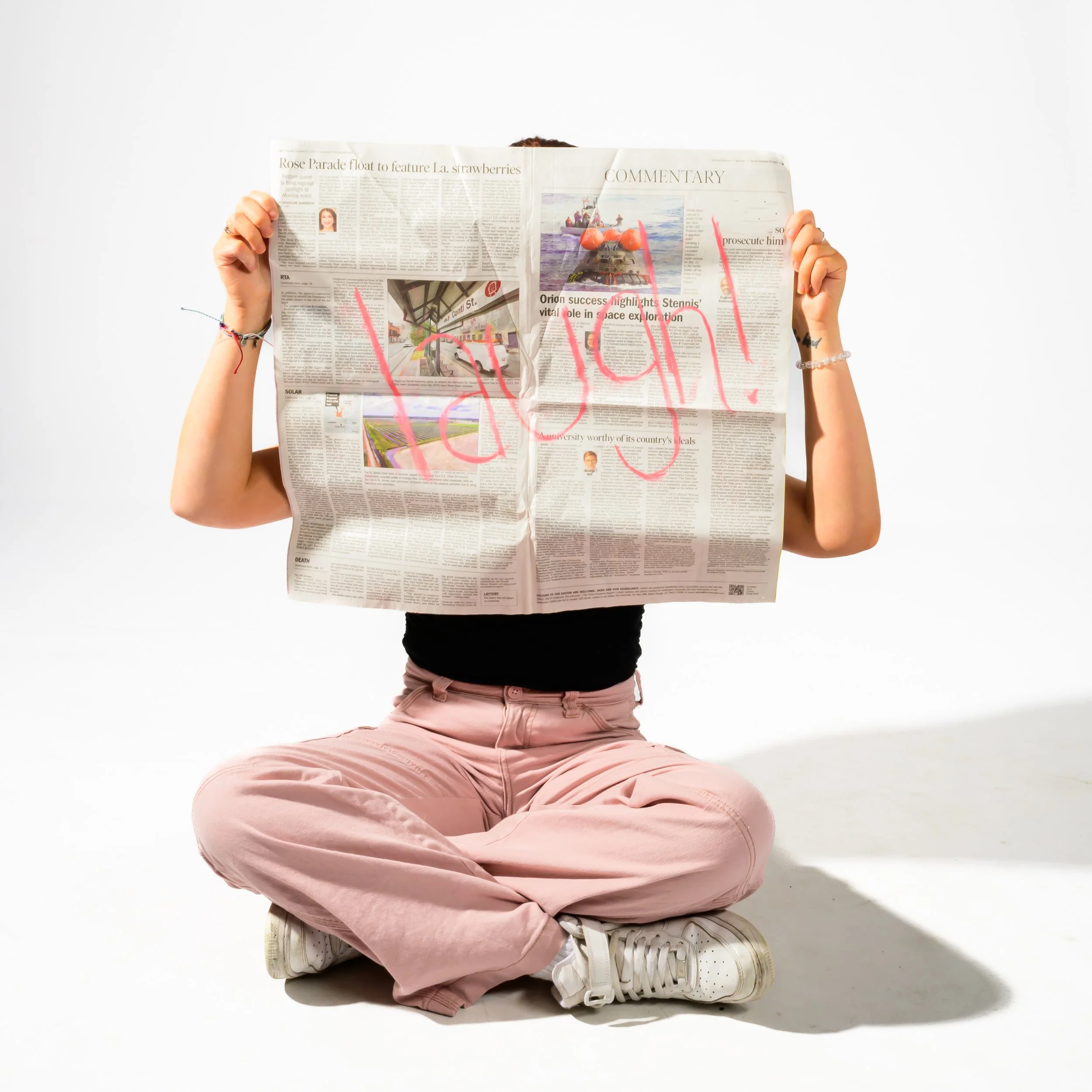 Person sitting cross-legged on the floor holding a newspaper with the word 'happy' written on it in pink marker, against a plain white background.